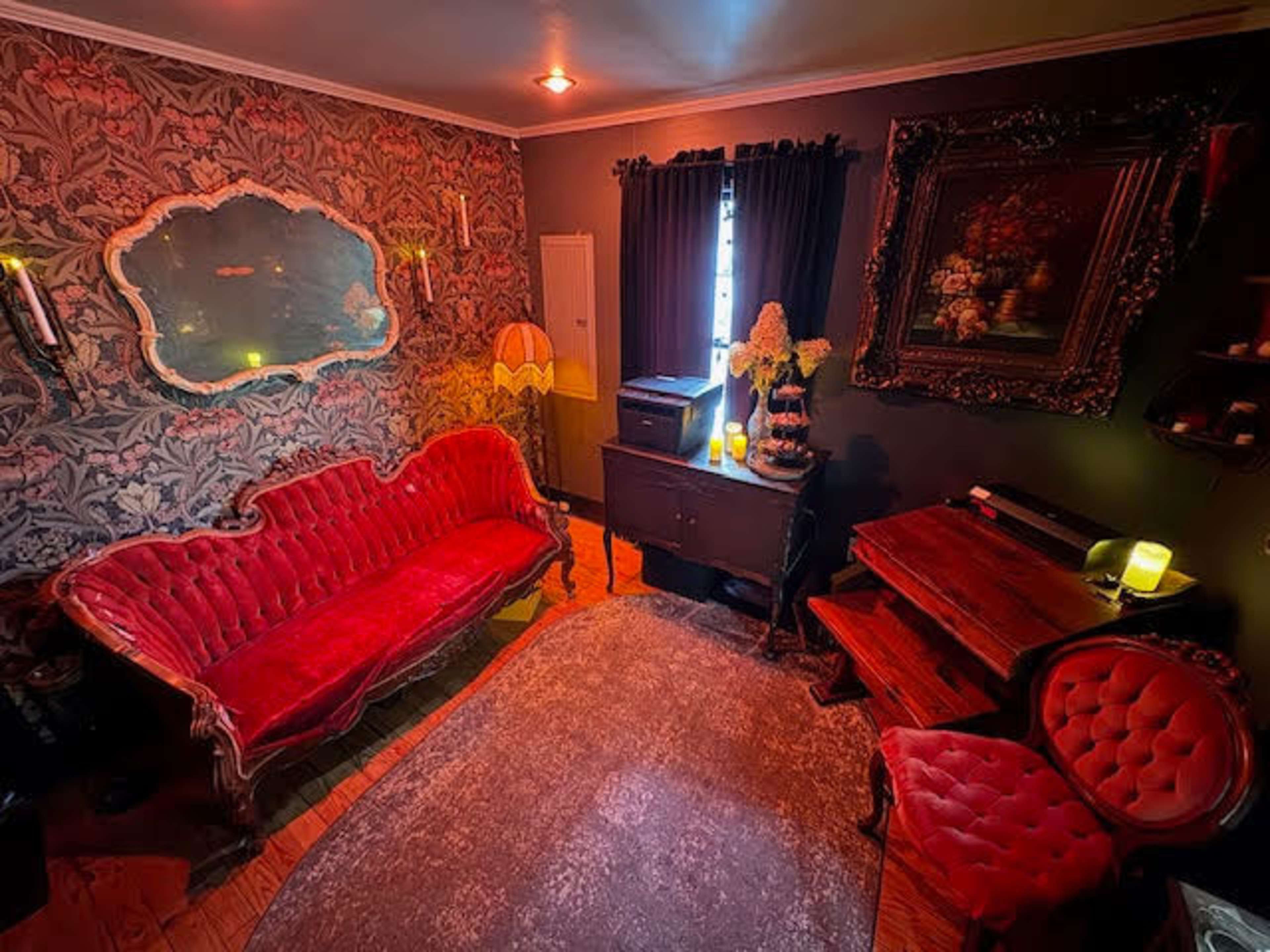 A vintage-inspired room features a red velvet sofa, a dark wooden dresser, and ornate decor against a patterned wallpaper backdrop.
