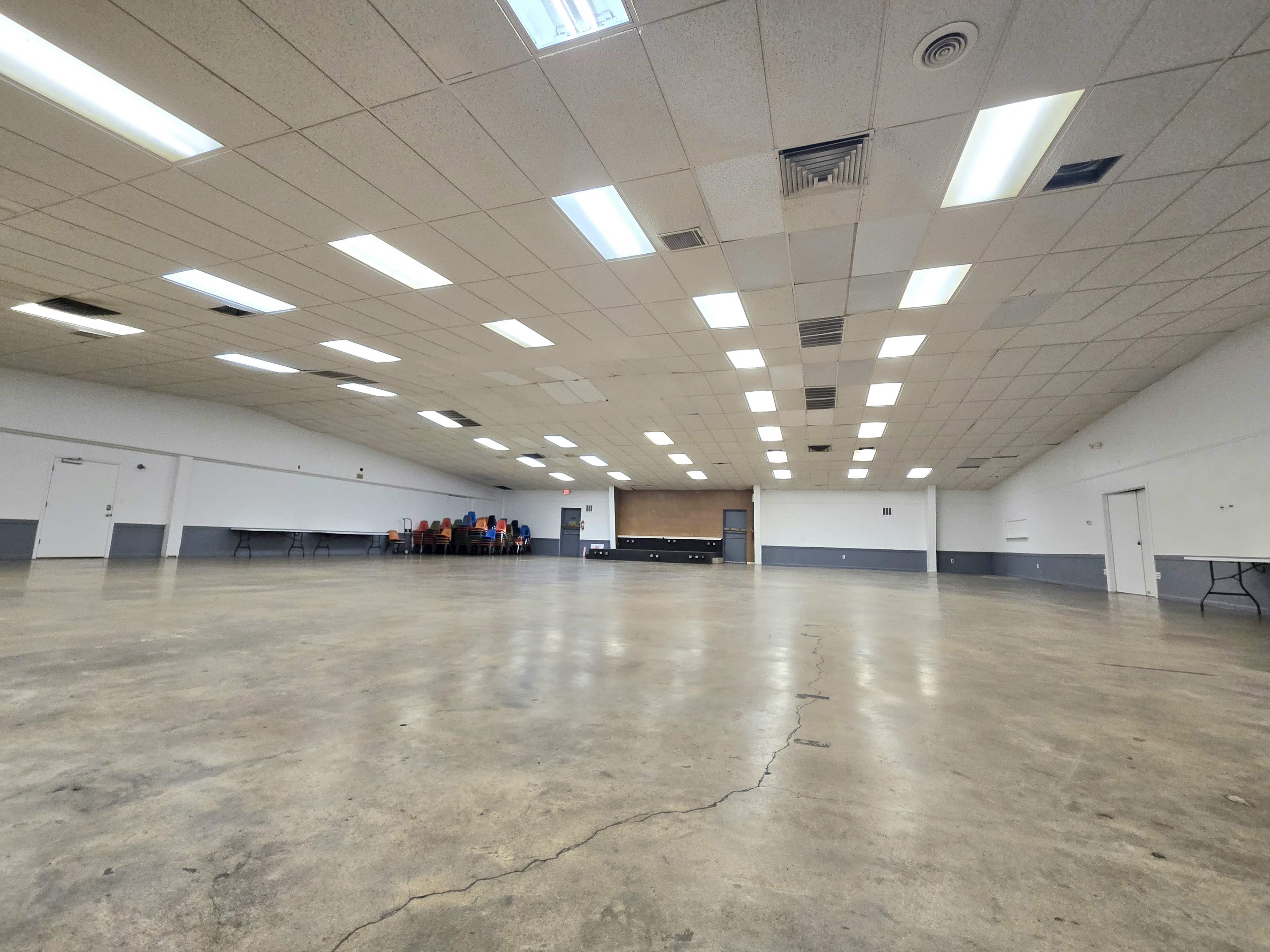 The image shows a large, empty hall with a polished concrete floor and rows of tables and chairs aligned against the walls.