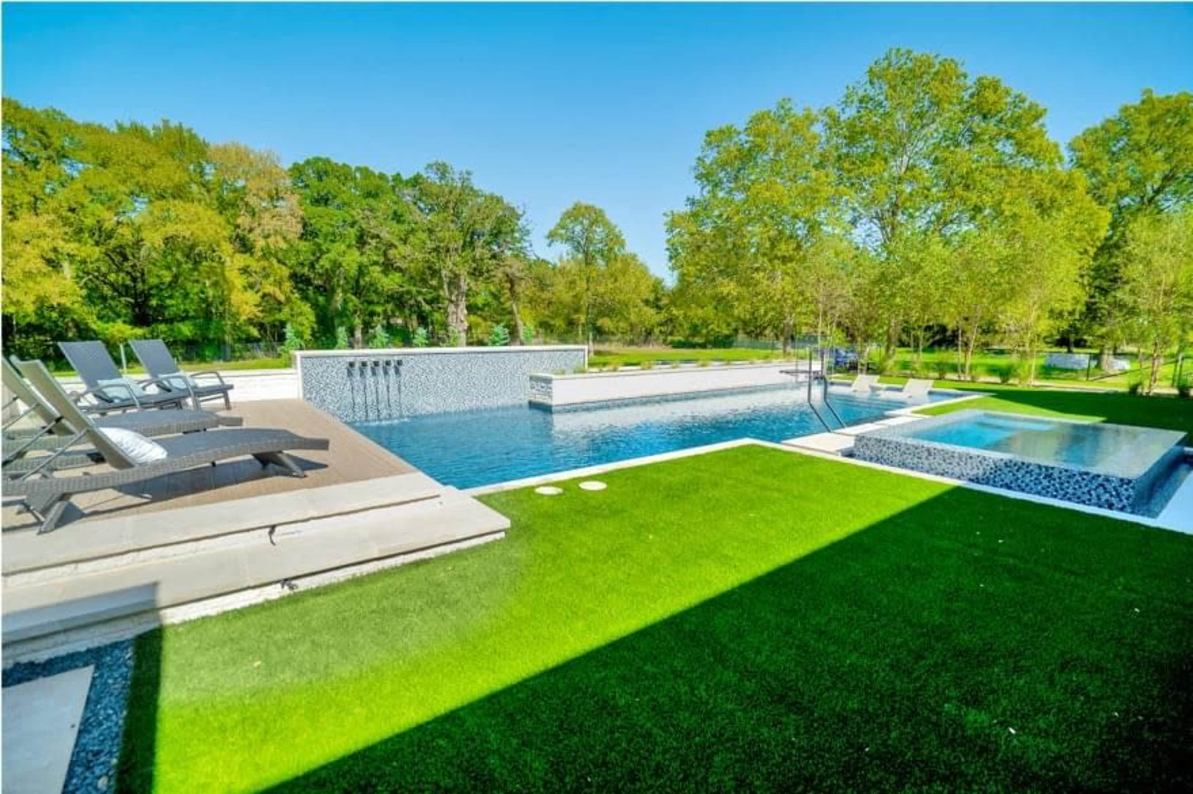 The image shows a modern swimming pool surrounded by greenery, with lounging chairs and a hot tub nearby.