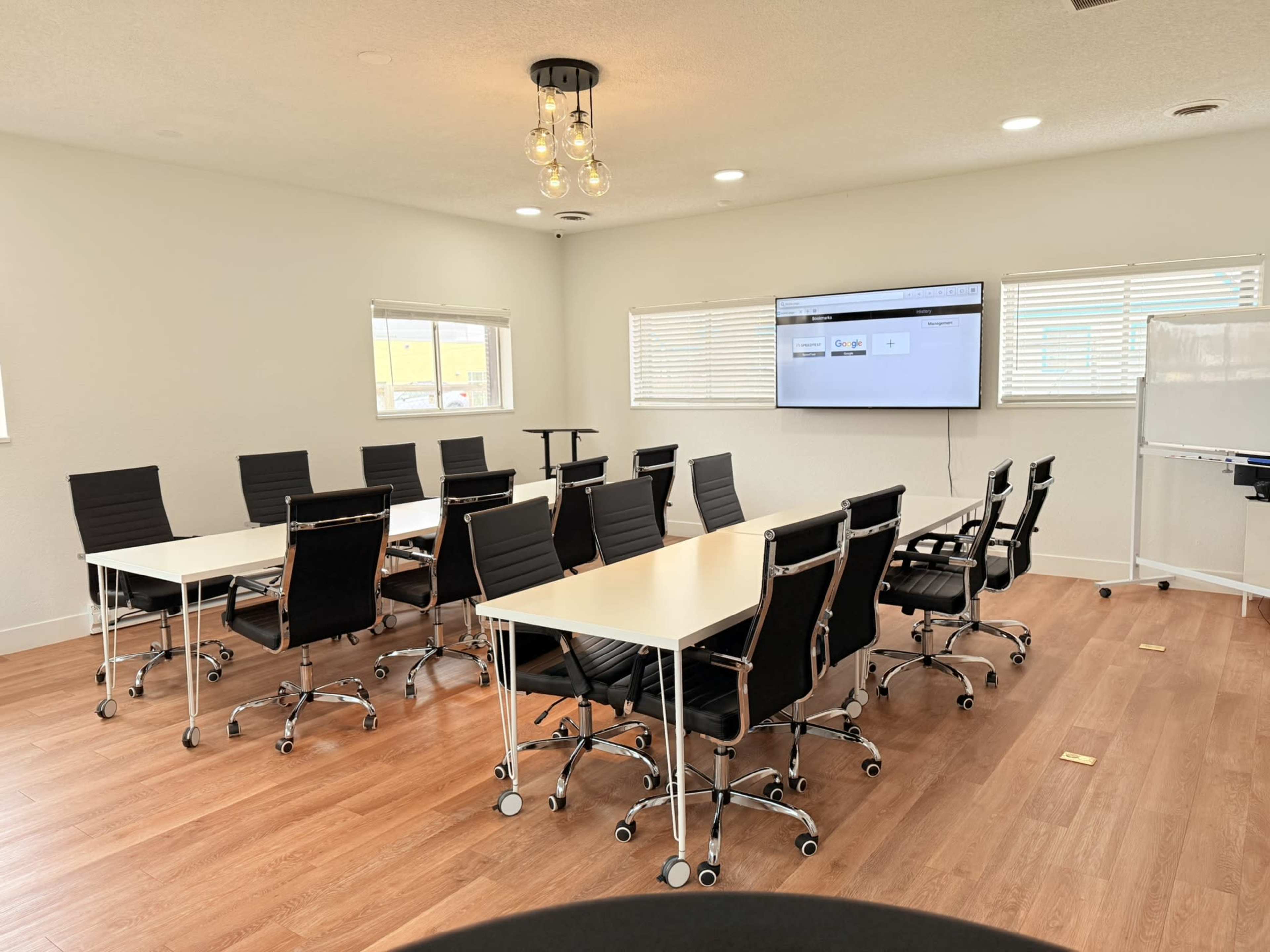 The image depicts a modern conference room with multiple white tables arranged in a U-shape, surrounded by black office chairs, and featuring a large screen on the wall.
