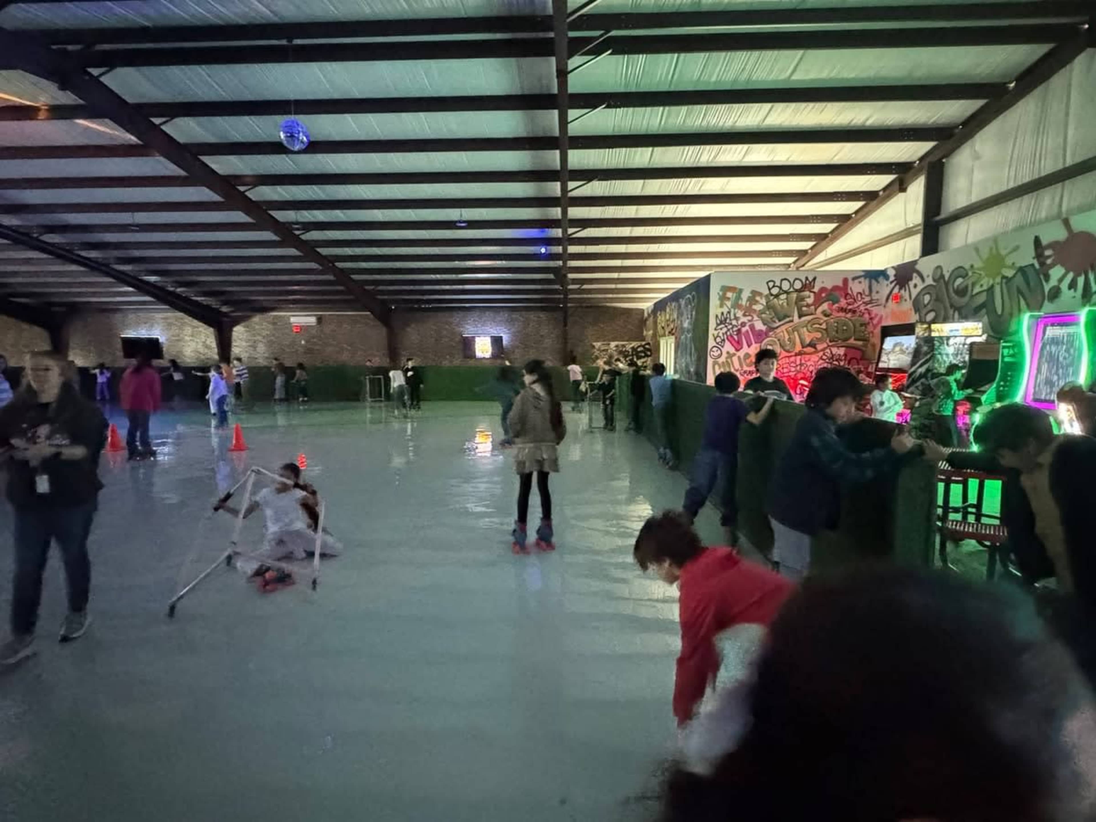 The image shows a roller skating rink with people skating and playing games, illuminated by colorful lights and decorated with graffiti along the walls.