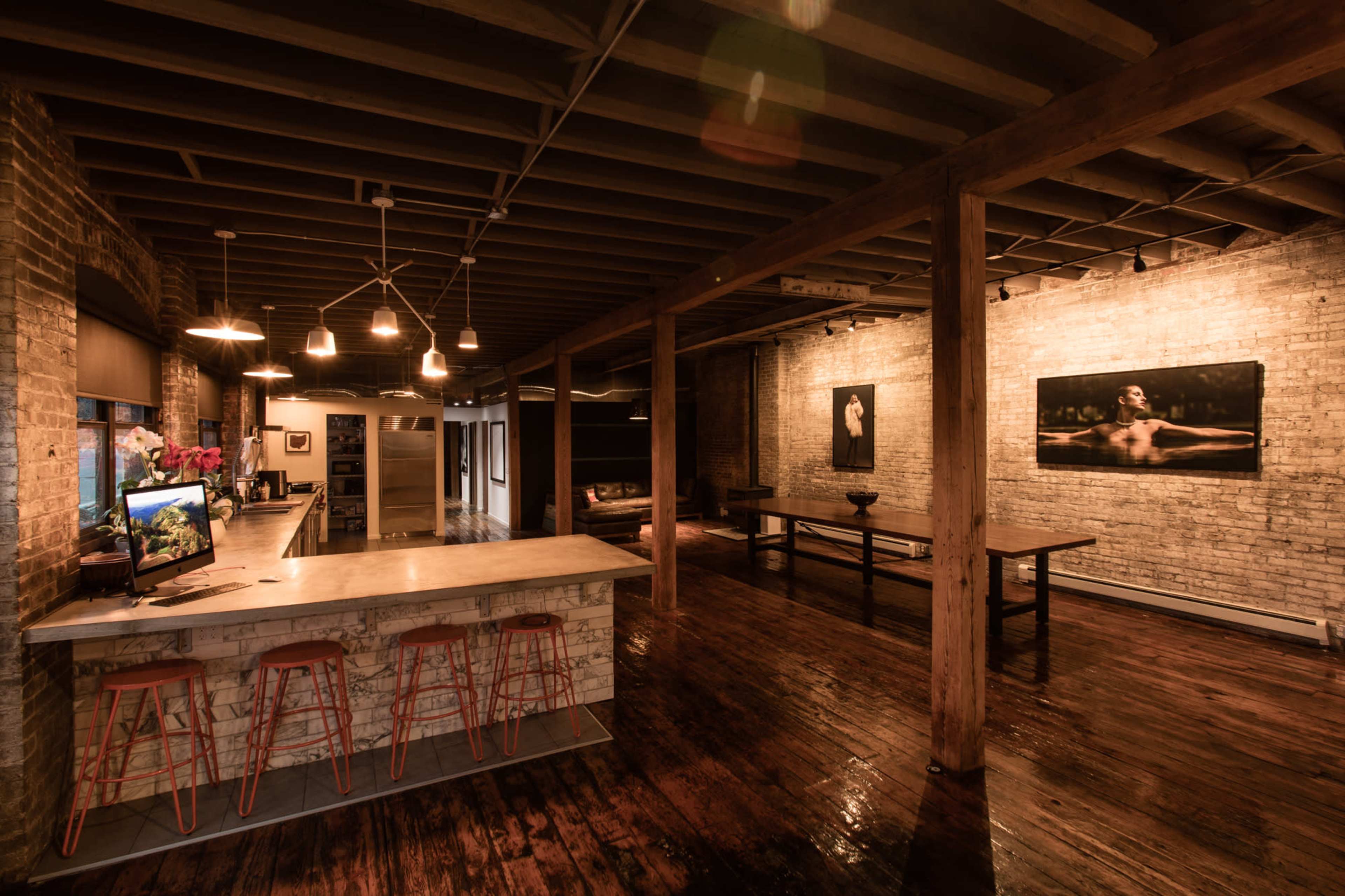 The image shows an open-concept interior space with exposed brick walls, wooden beams, and a kitchen island with bar stools.