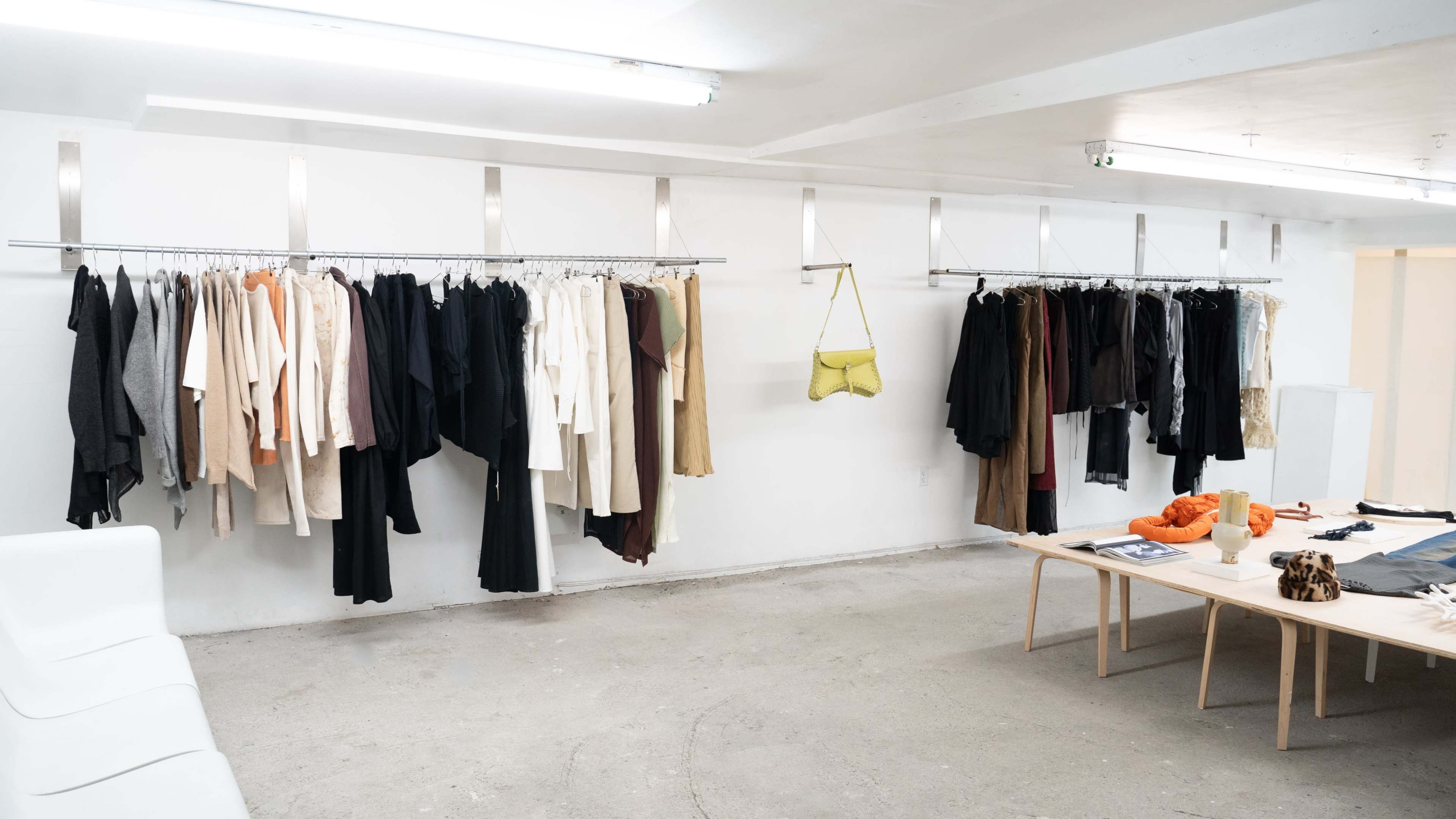 A minimalist clothing display featuring various garments on racks along a white wall, with a table set with accessories and a few items.