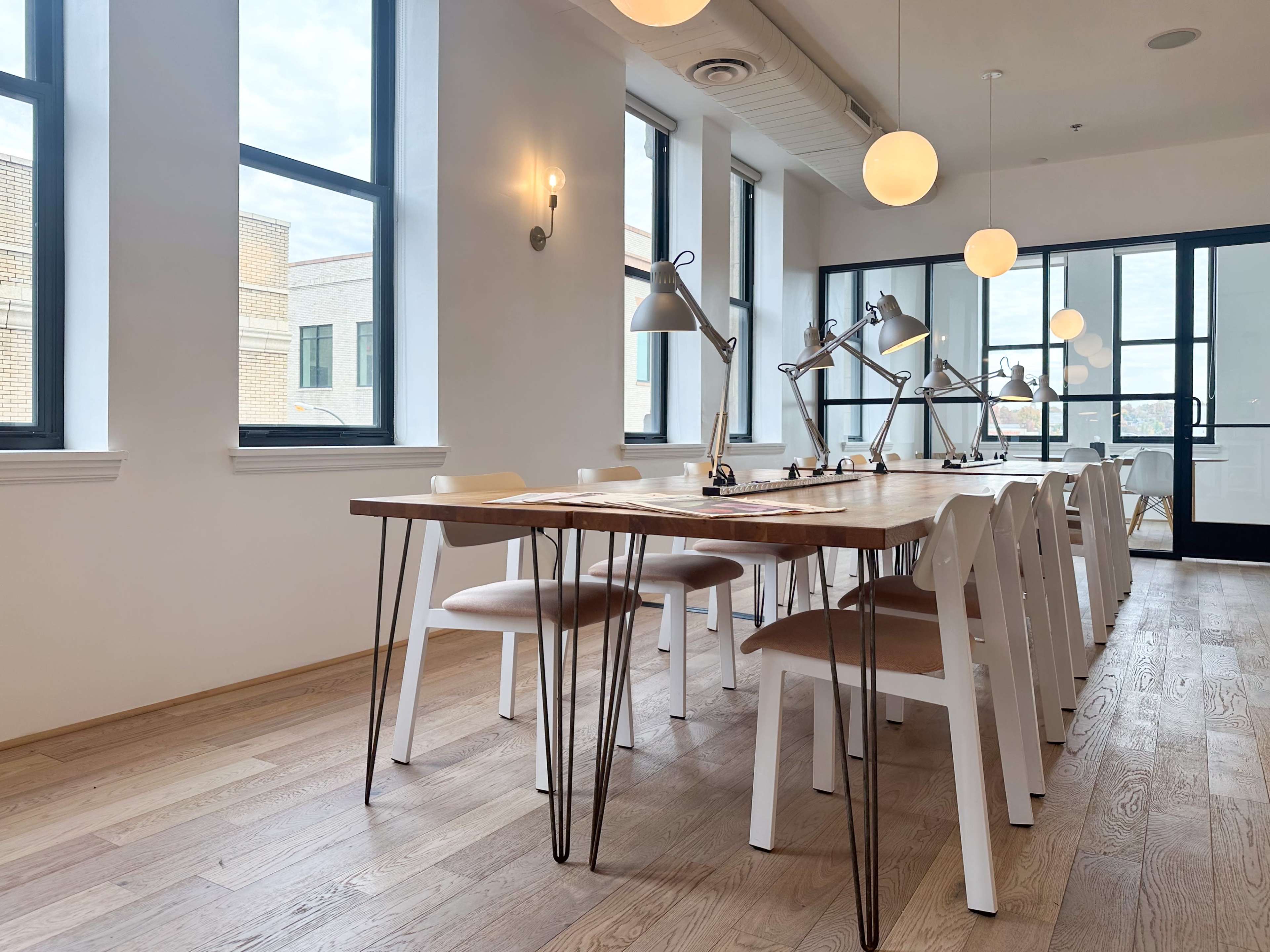 A modern conference room features a long wooden table surrounded by eight white chairs and several desk lamps, with large windows allowing natural light to enter.