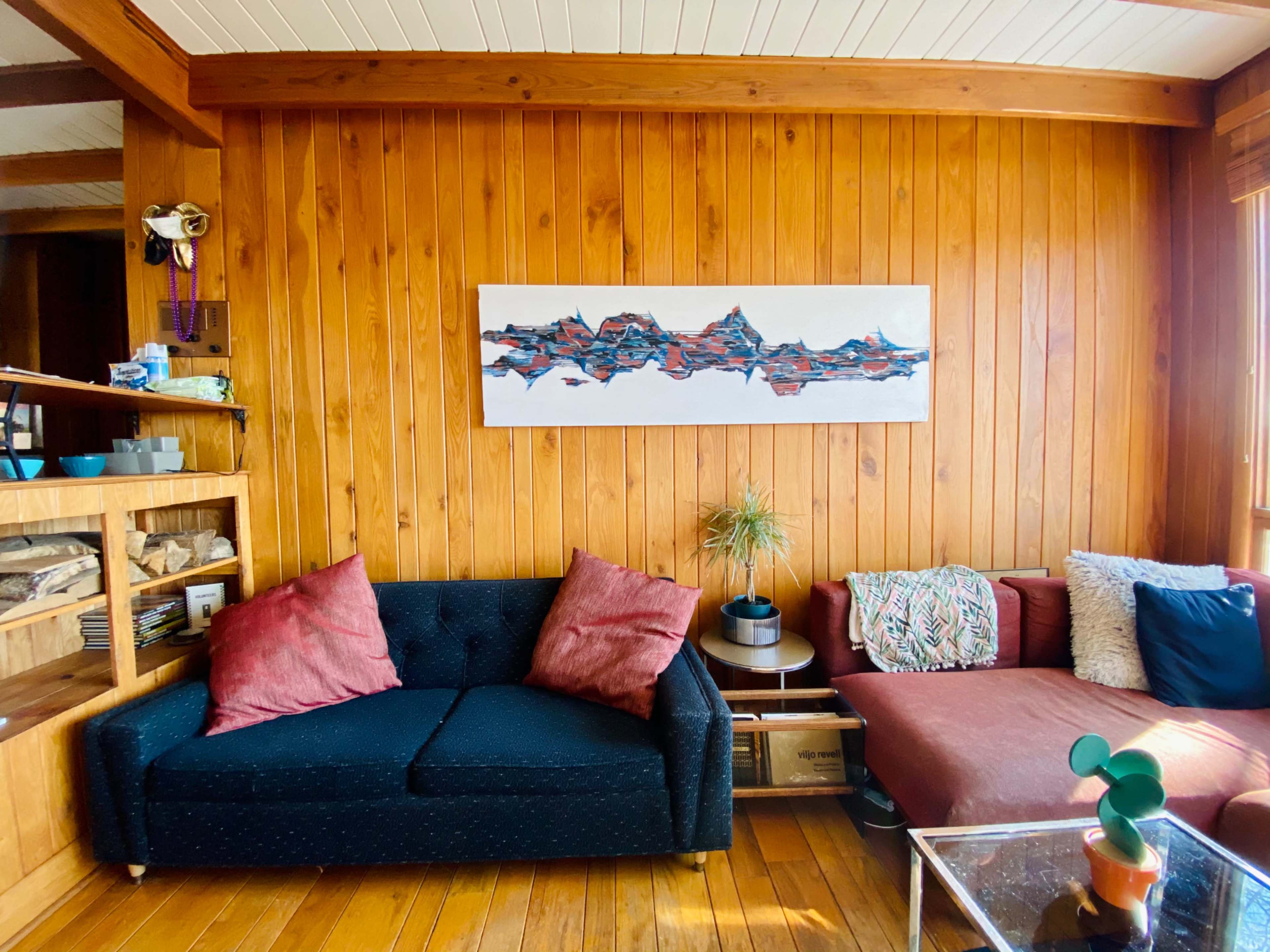 A room with wooden walls featuring two sofas, one dark with red pillows and another in a light color, along with a shelf of items and a large artwork on the wall.