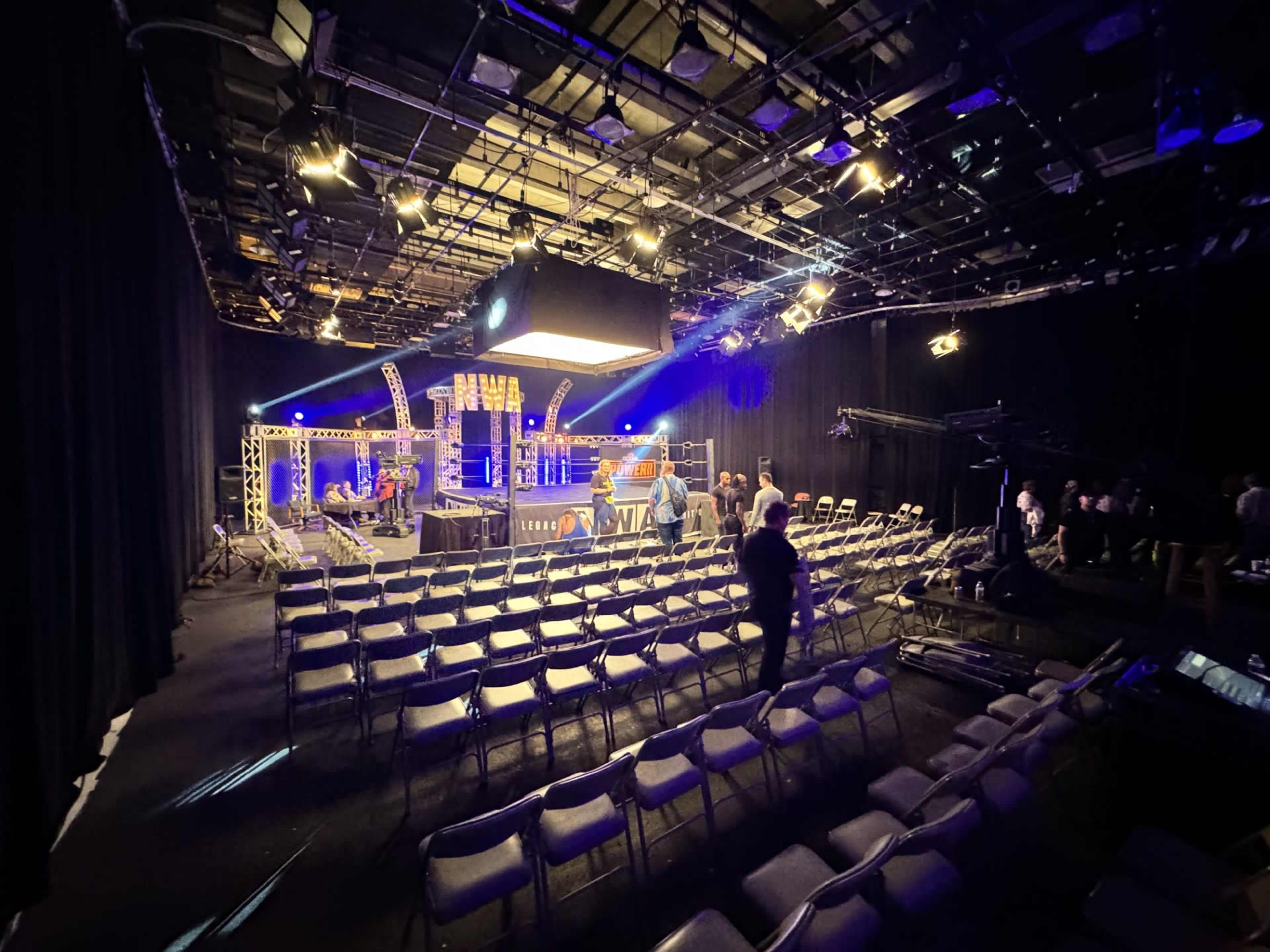 A studio setting with rows of empty chairs facing a well-lit stage, equipped with lighting and camera gear.