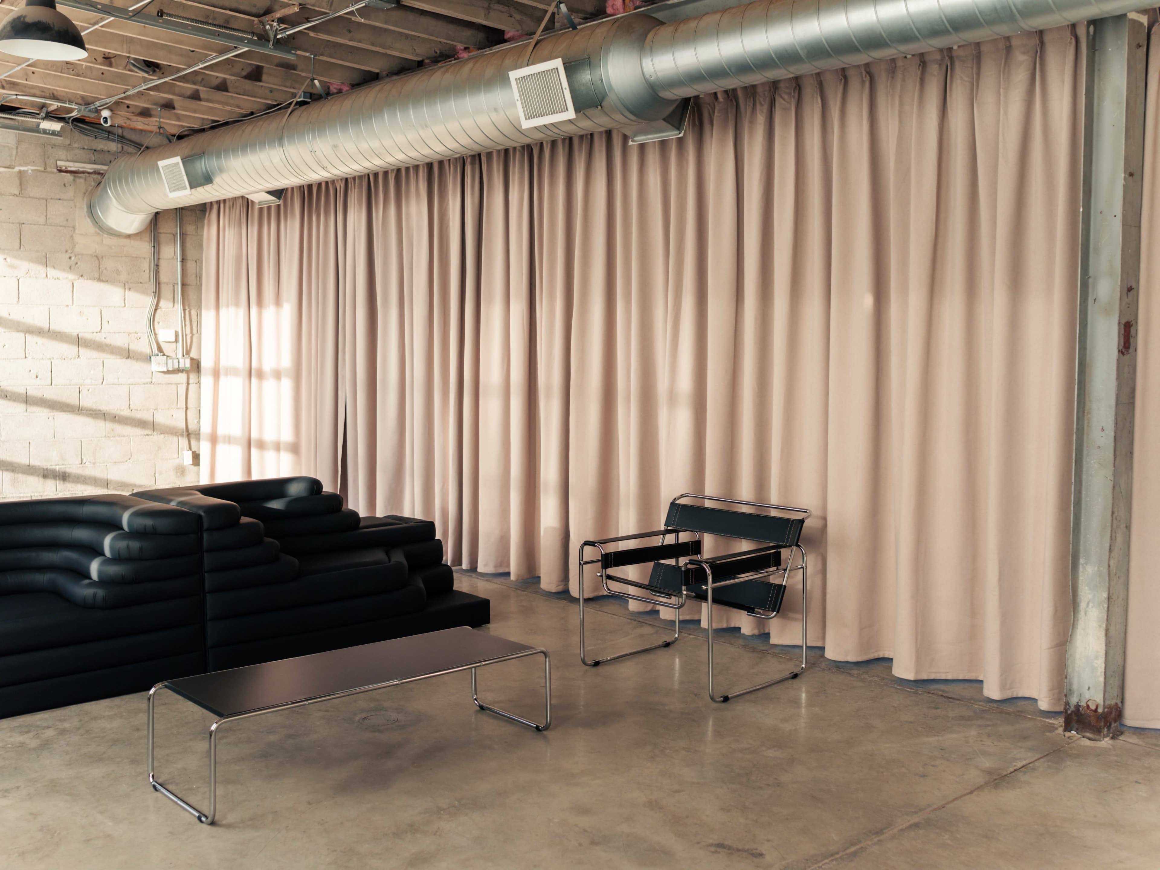 The image shows a minimalistic interior featuring a black lounge chair and a low coffee table in front of a wall of beige curtains.