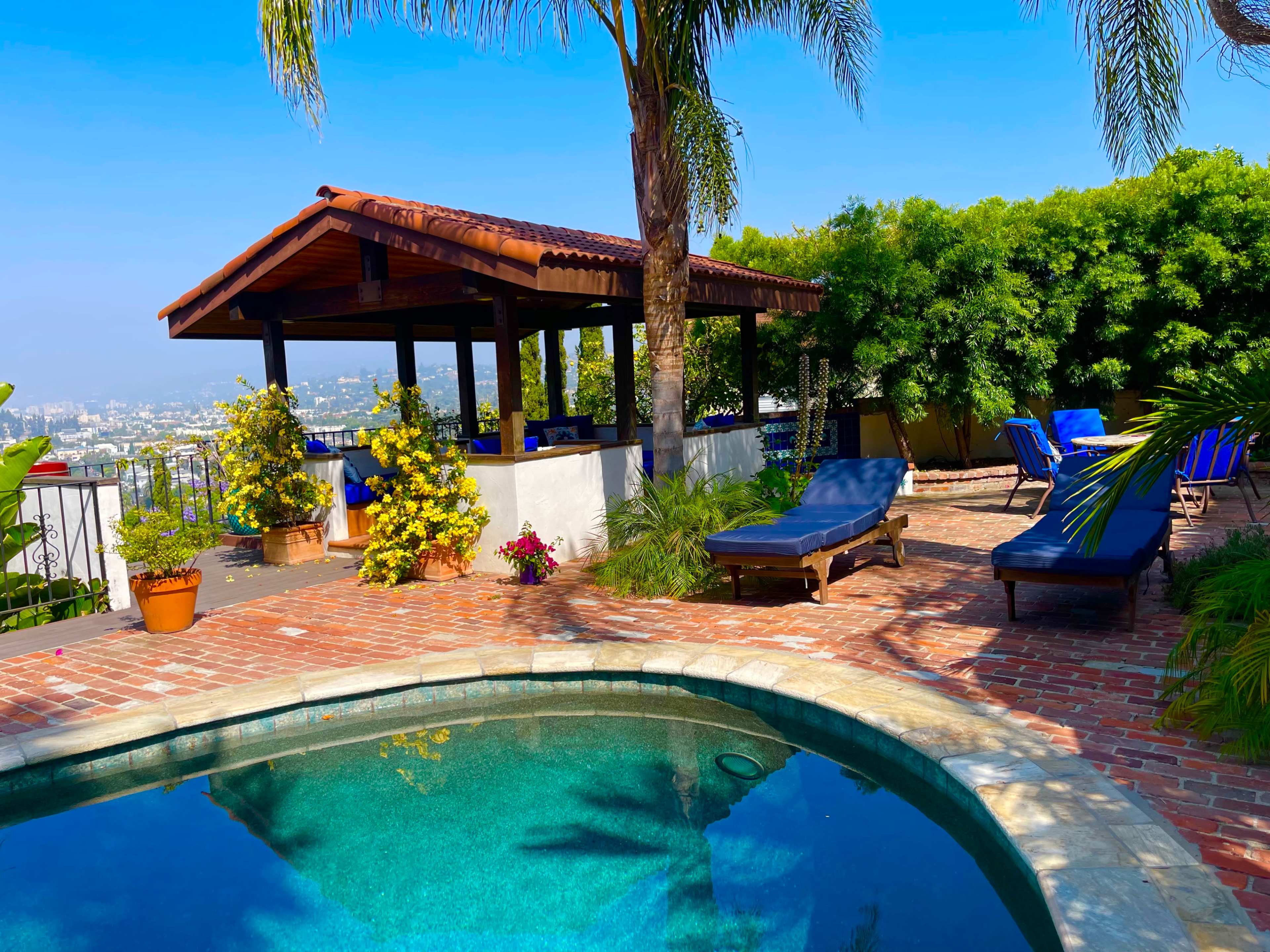 A backyard features a swimming pool, lounge chairs, a shaded gazebo, and potted plants with a view of the city in the background.