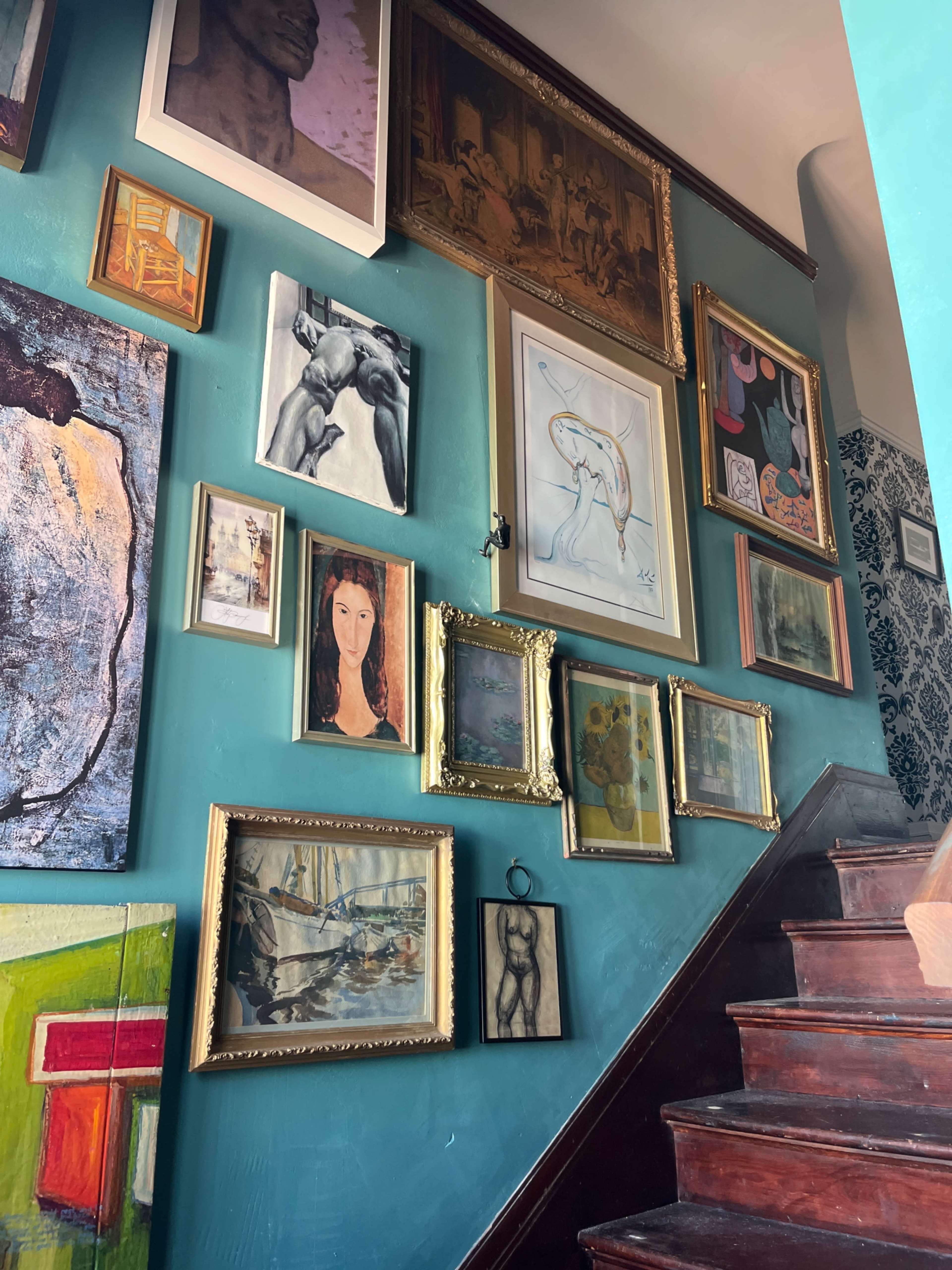 The image shows a staircase wall adorned with a diverse collection of framed artworks, including paintings and drawings in various styles, arranged visually along the teal-painted surface.