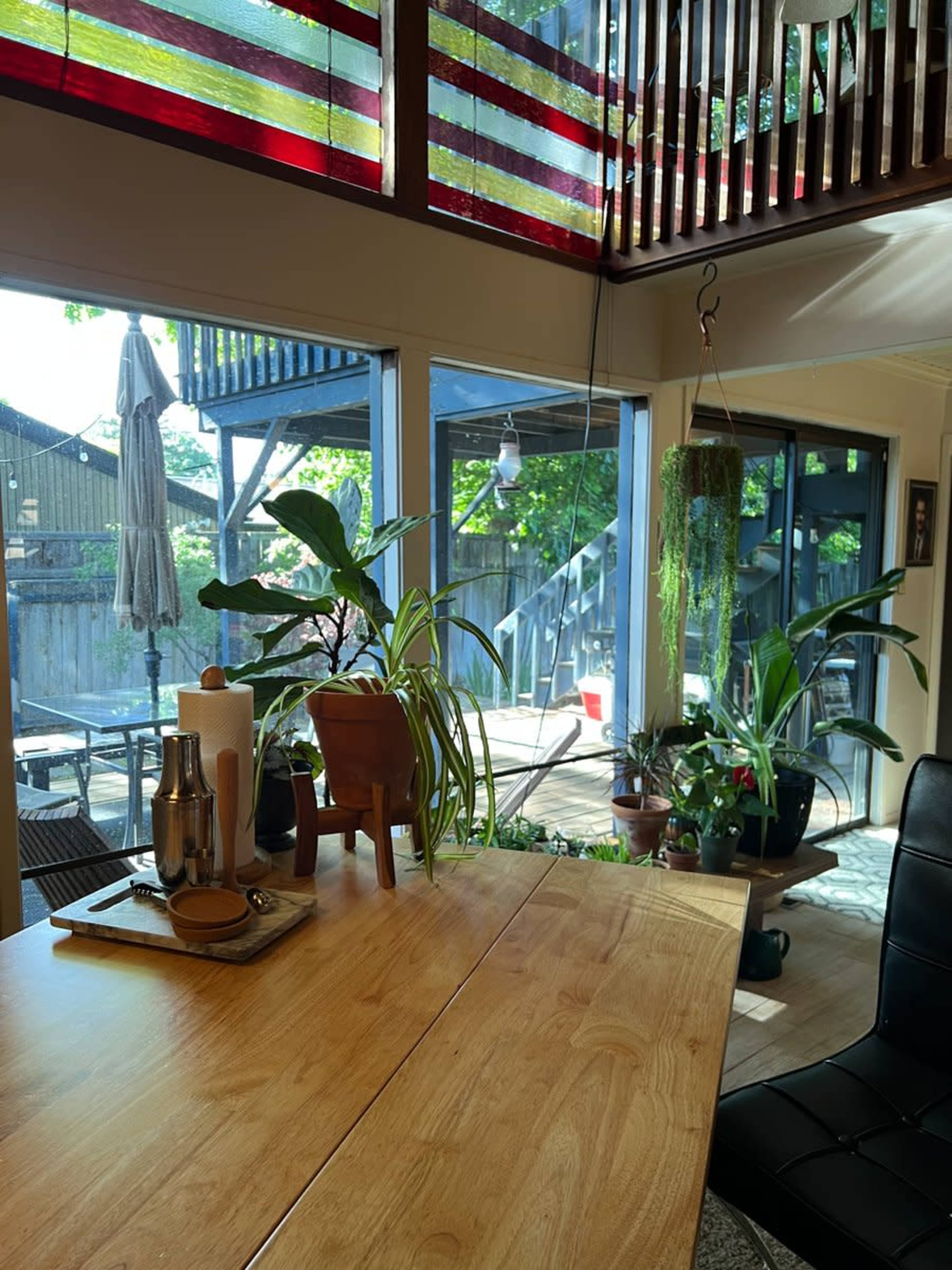 A wooden dining table with a tray sits in a room filled with plants, overlooking a deck with outdoor furniture through large glass doors.