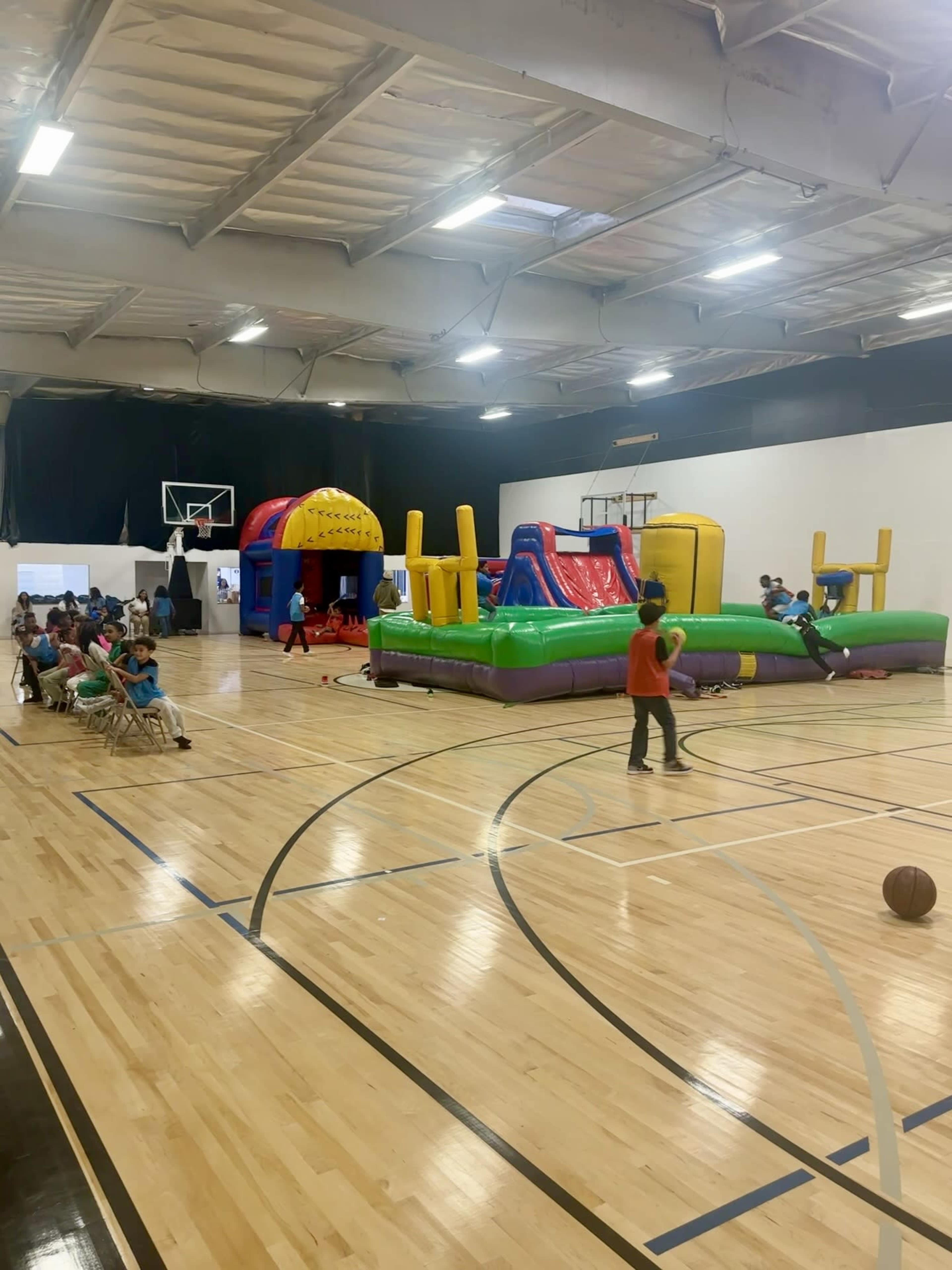 A large indoor gymnasium features several inflatable bounce houses and slides, with children playing and sitting on chairs in the background.