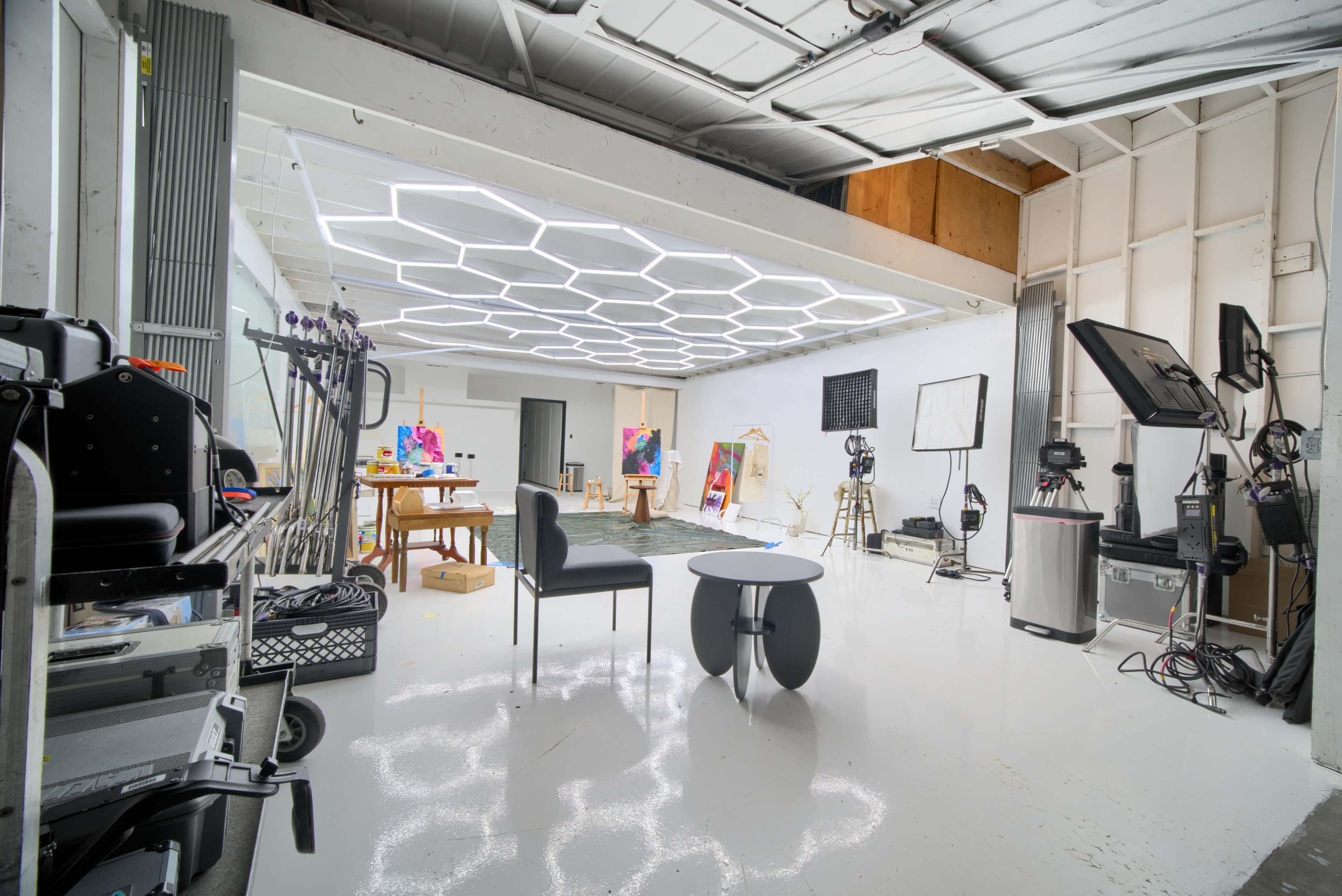 The image shows a spacious, well-lit studio with a modern design, featuring a hexagonal light installation, art supplies, and equipment arranged for creative work.