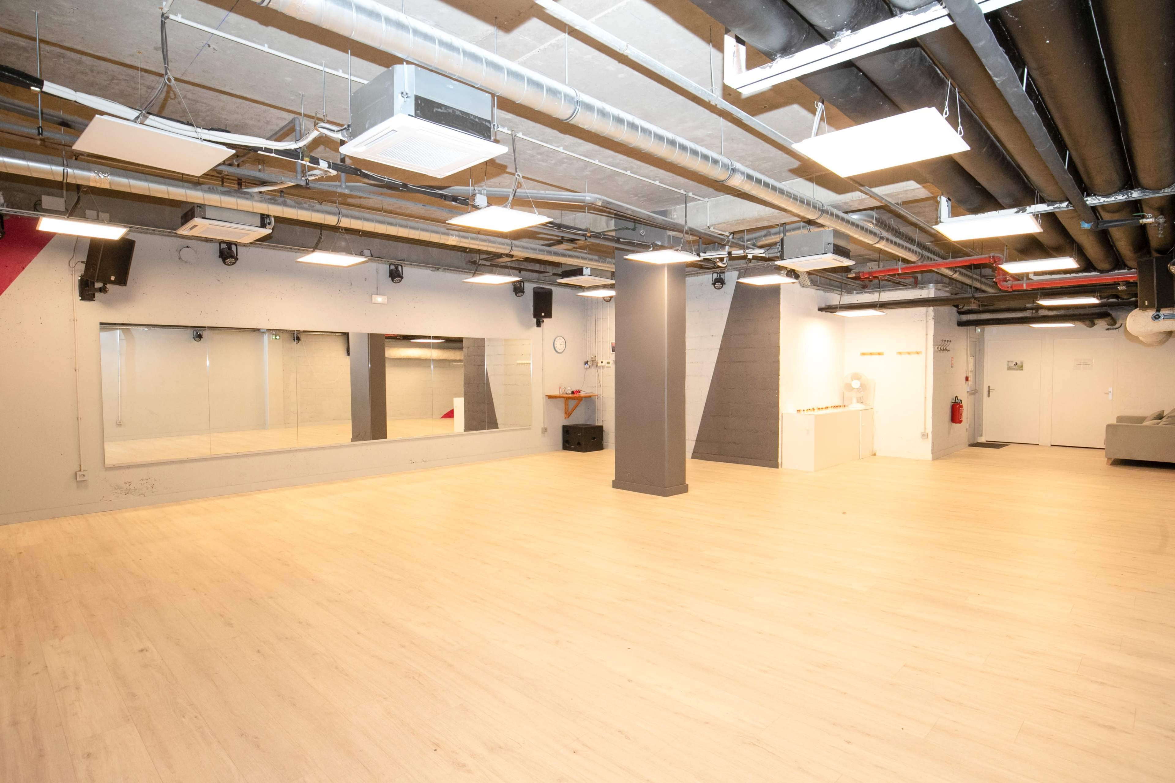 A spacious, well-lit studio with wooden flooring, mirrors on one wall, and exposed piping on the ceiling.