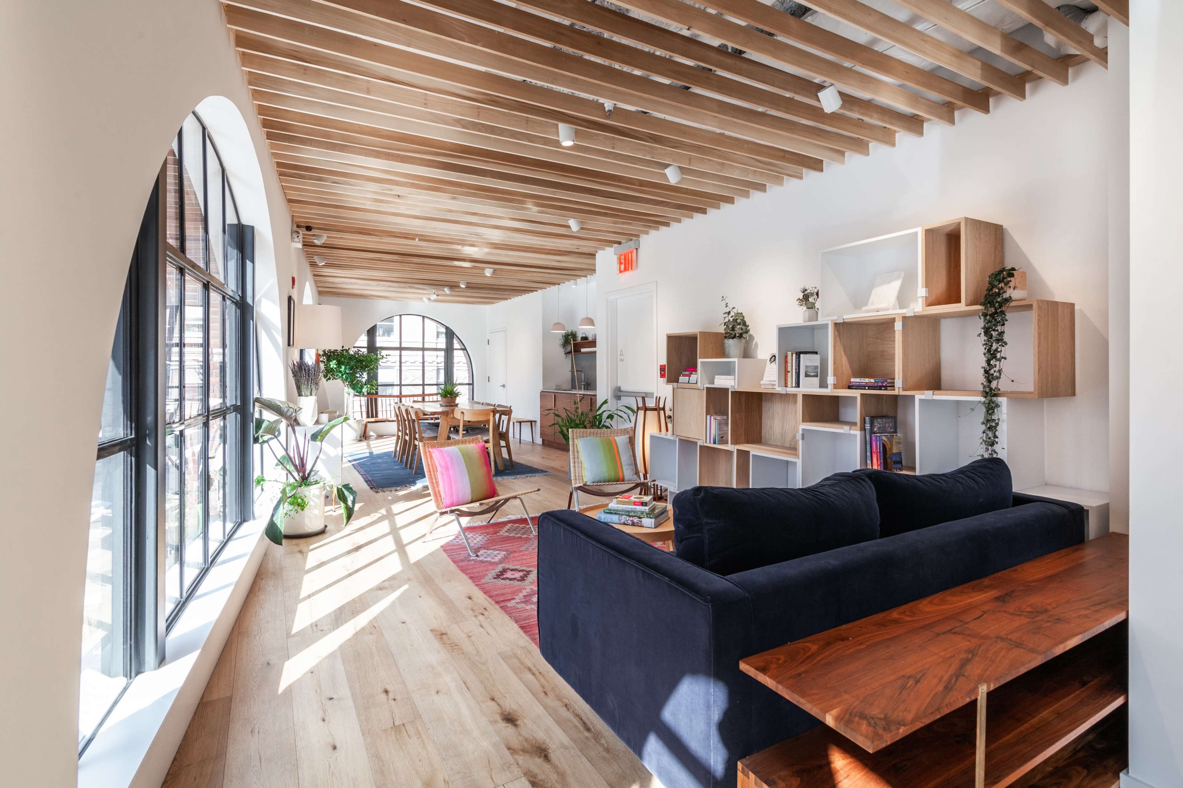 A bright and spacious interior with wooden ceiling beams, large windows, a dark blue sofa, a wooden table, and shelving filled with books and decorative items.