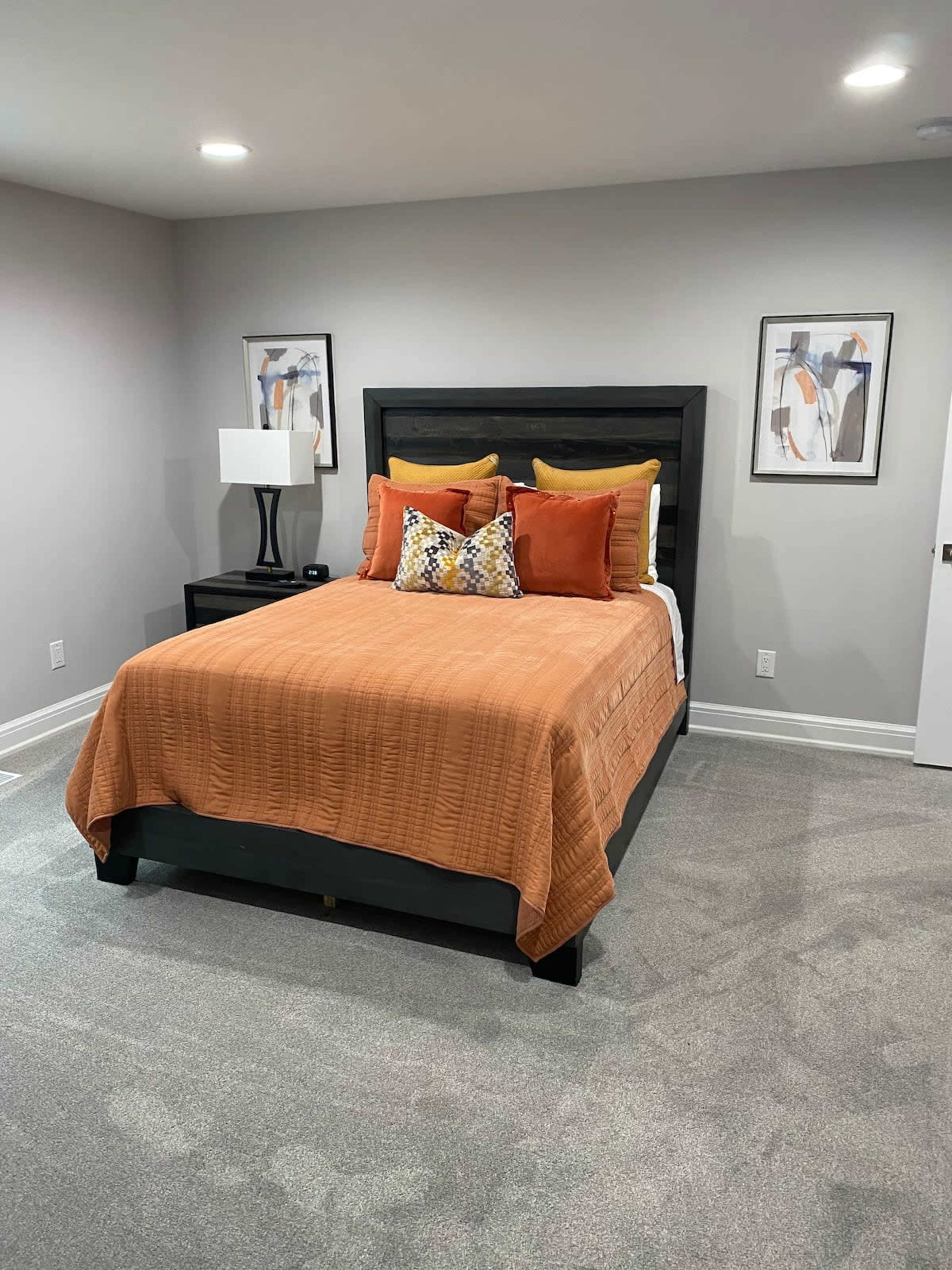 A neatly arranged bedroom with a black bed frame, an orange bedspread, and decorative pillows, along with a nightstand and wall art.