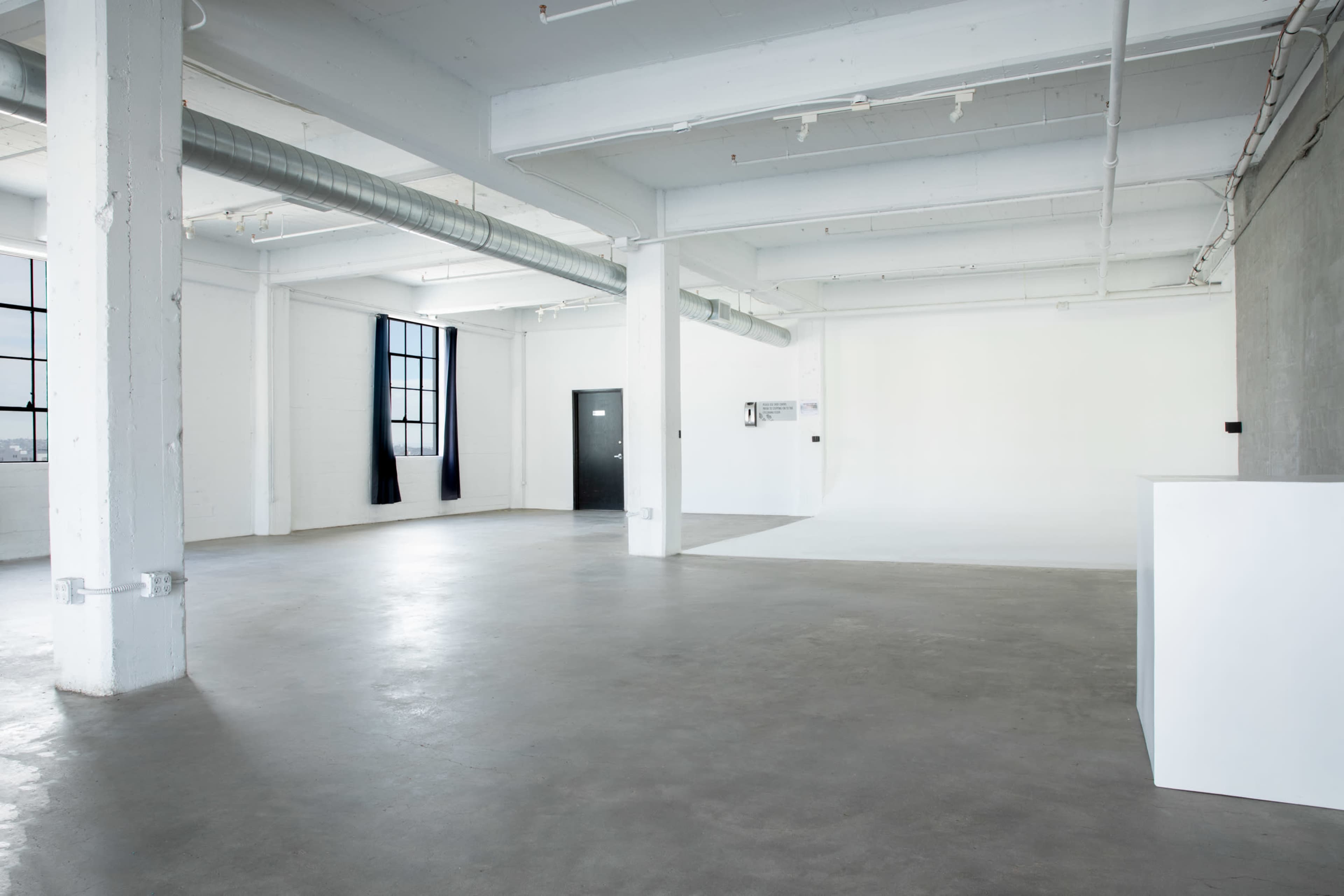 The image shows a spacious, empty industrial-style room with concrete floors, large windows, and minimal furnishings.