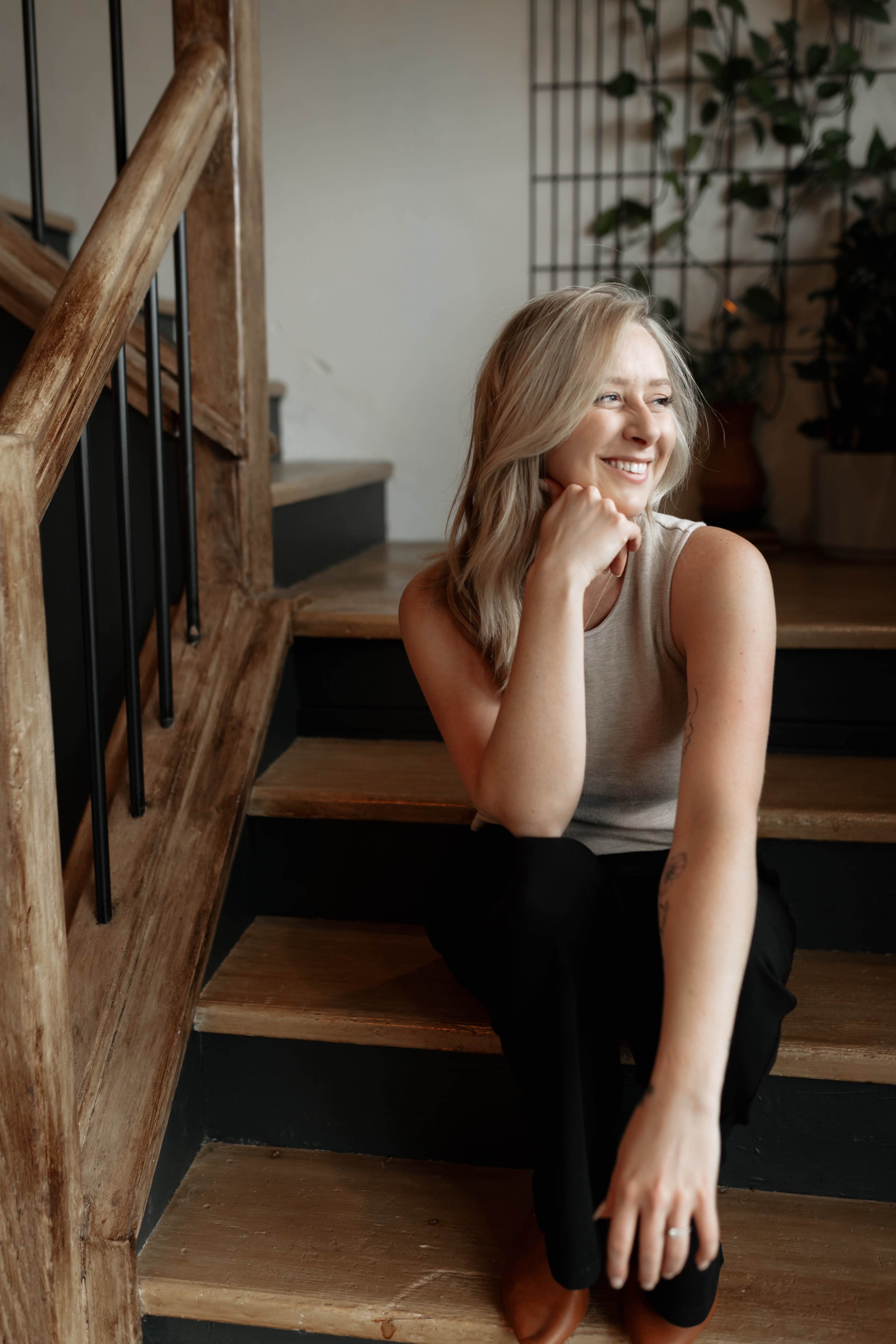 A person with long hair sits on a staircase, resting their chin on their hand and smiling.