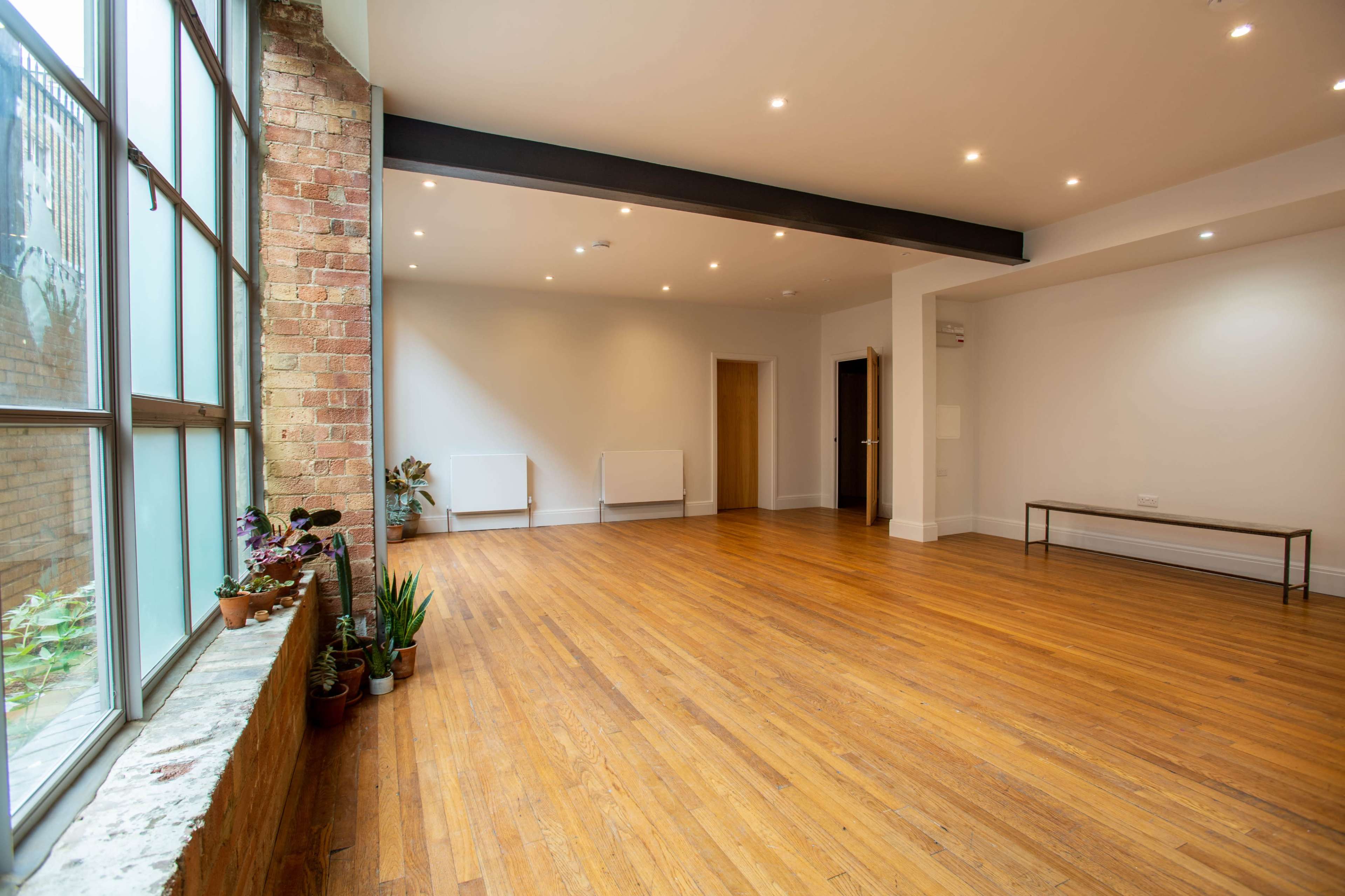 A spacious, empty room with wooden flooring and large windows, featuring minimal furniture and plants along the windowsill.