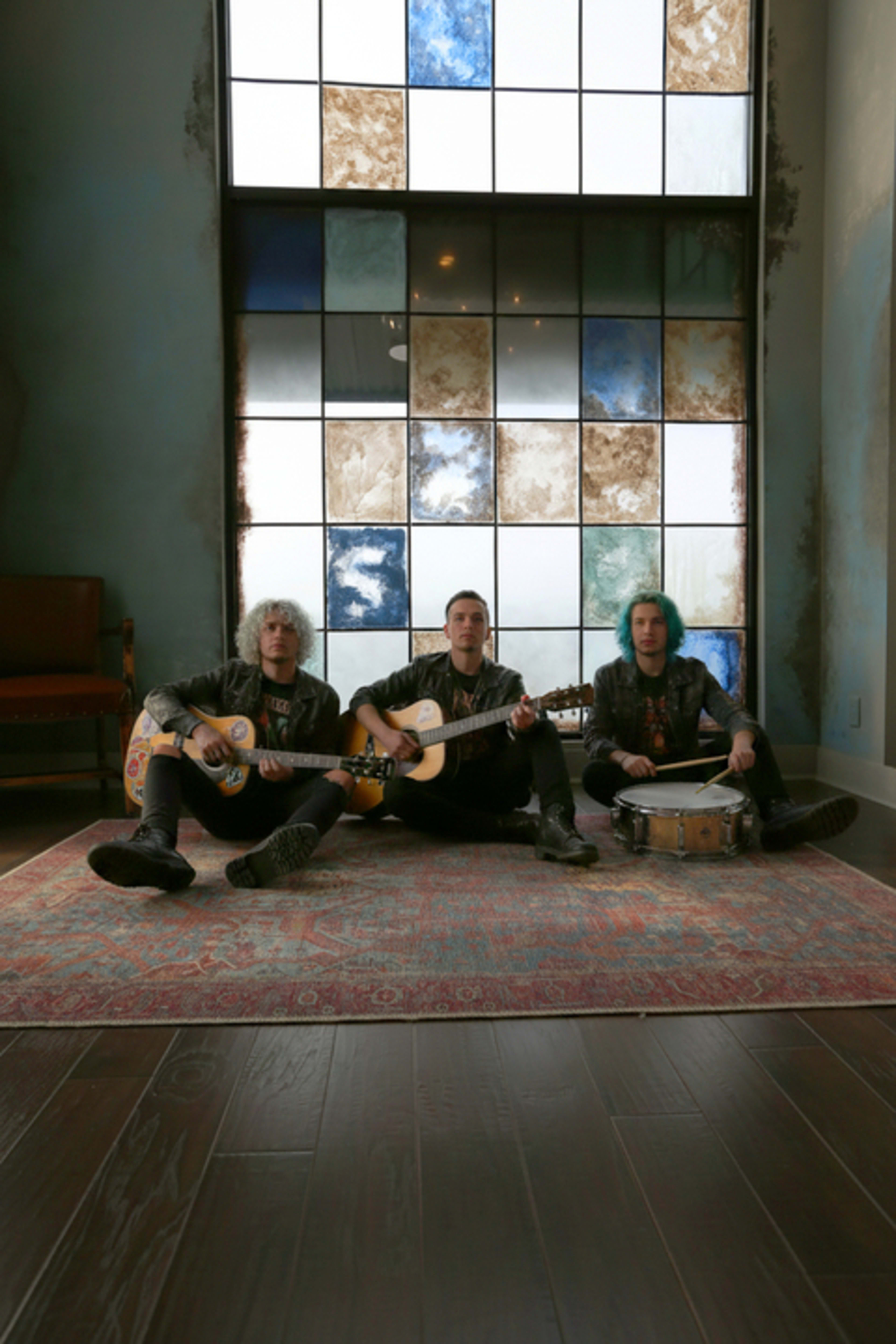 Three musicians sit on a patterned rug in a sunlit room with a large stained glass window behind them, each holding an instrument.