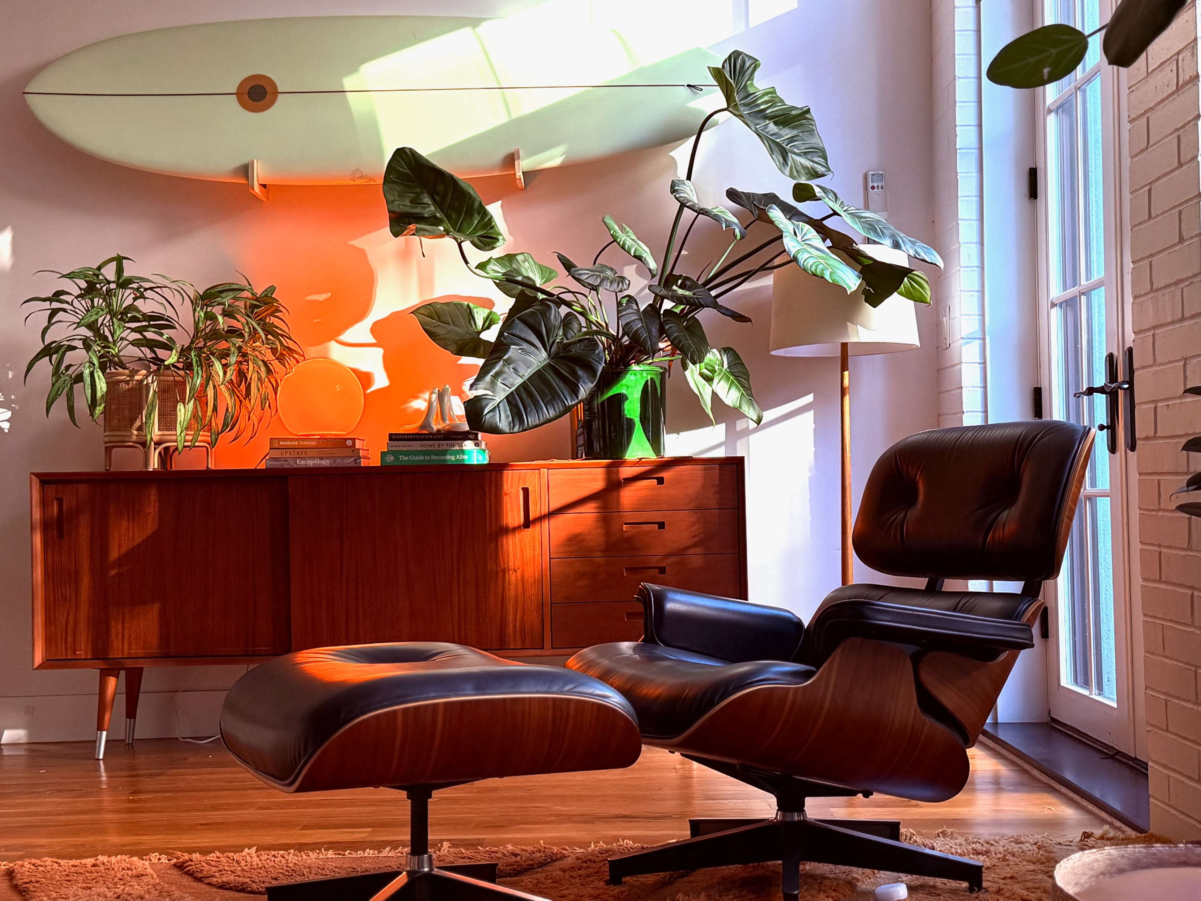 A modern living space features an Eames lounge chair and ottoman, surrounded by indoor plants and a wooden sideboard, with a surfboard mounted on the wall above.