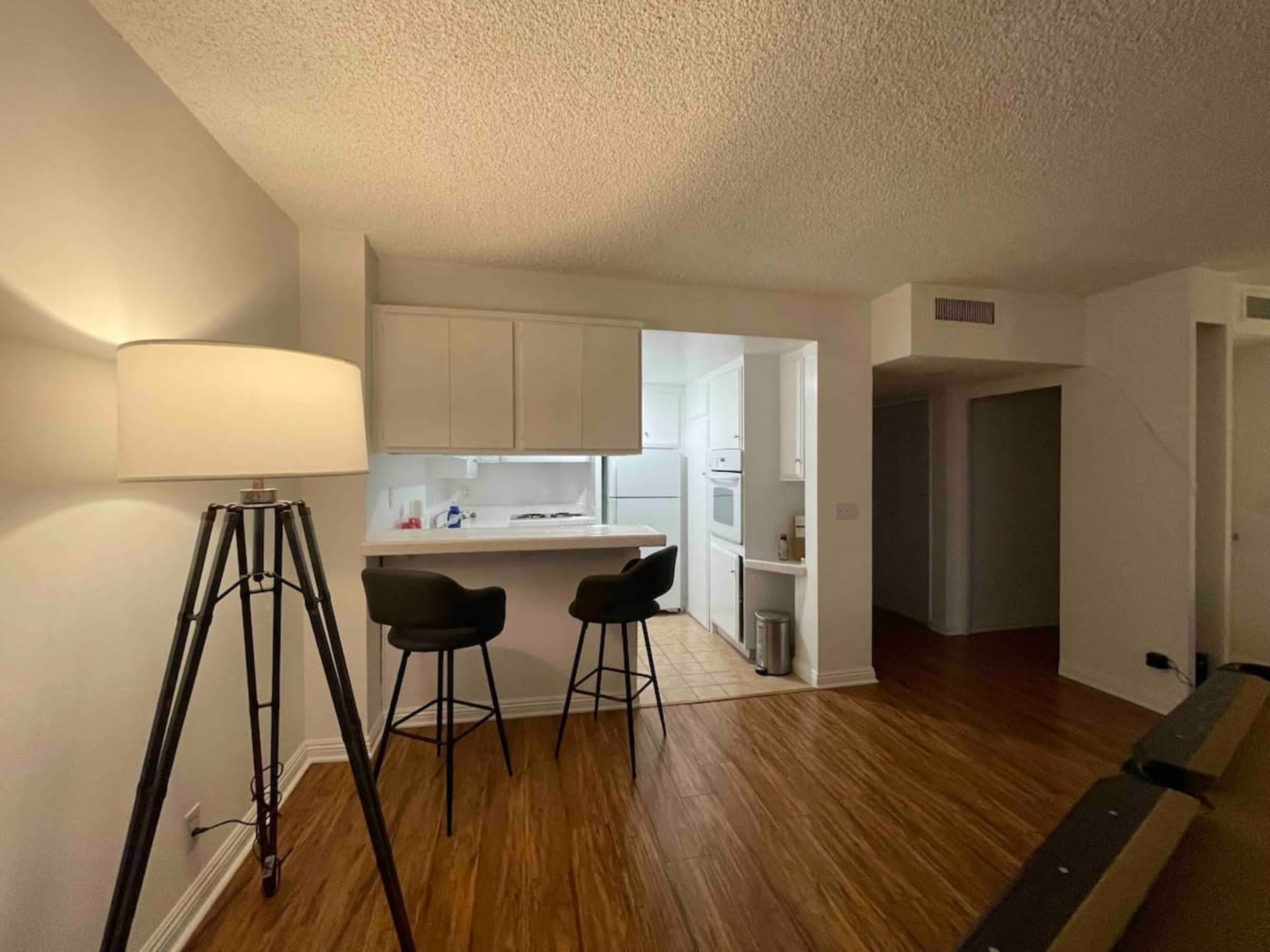 A small, modern kitchen with white cabinetry, a light-colored dining area featuring two stools, and a lamp in the foreground casting light.