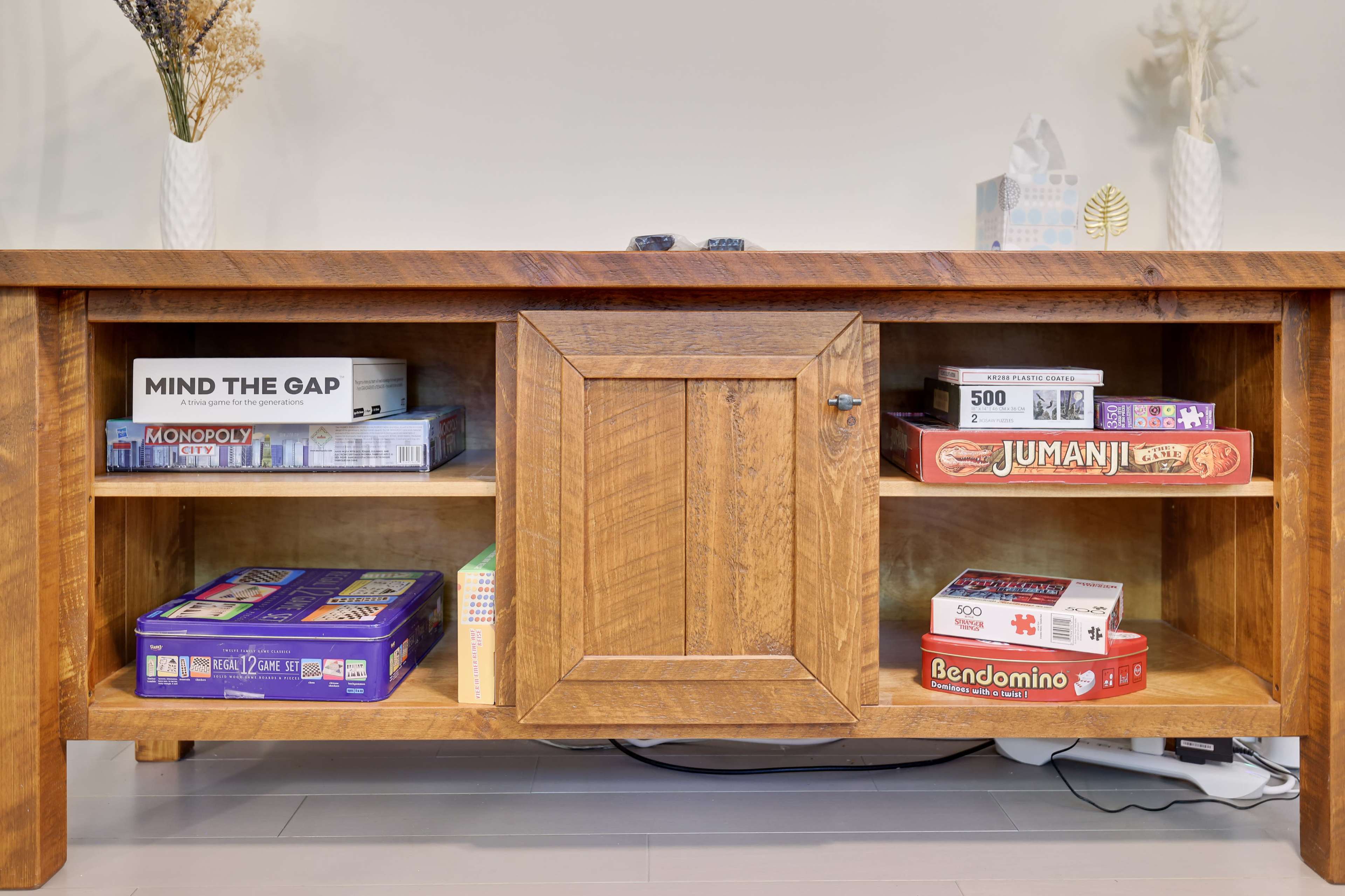 A wooden cabinet with an open section displaying various board games, including "Monopoly," "Jumanji," and "Bendomino."