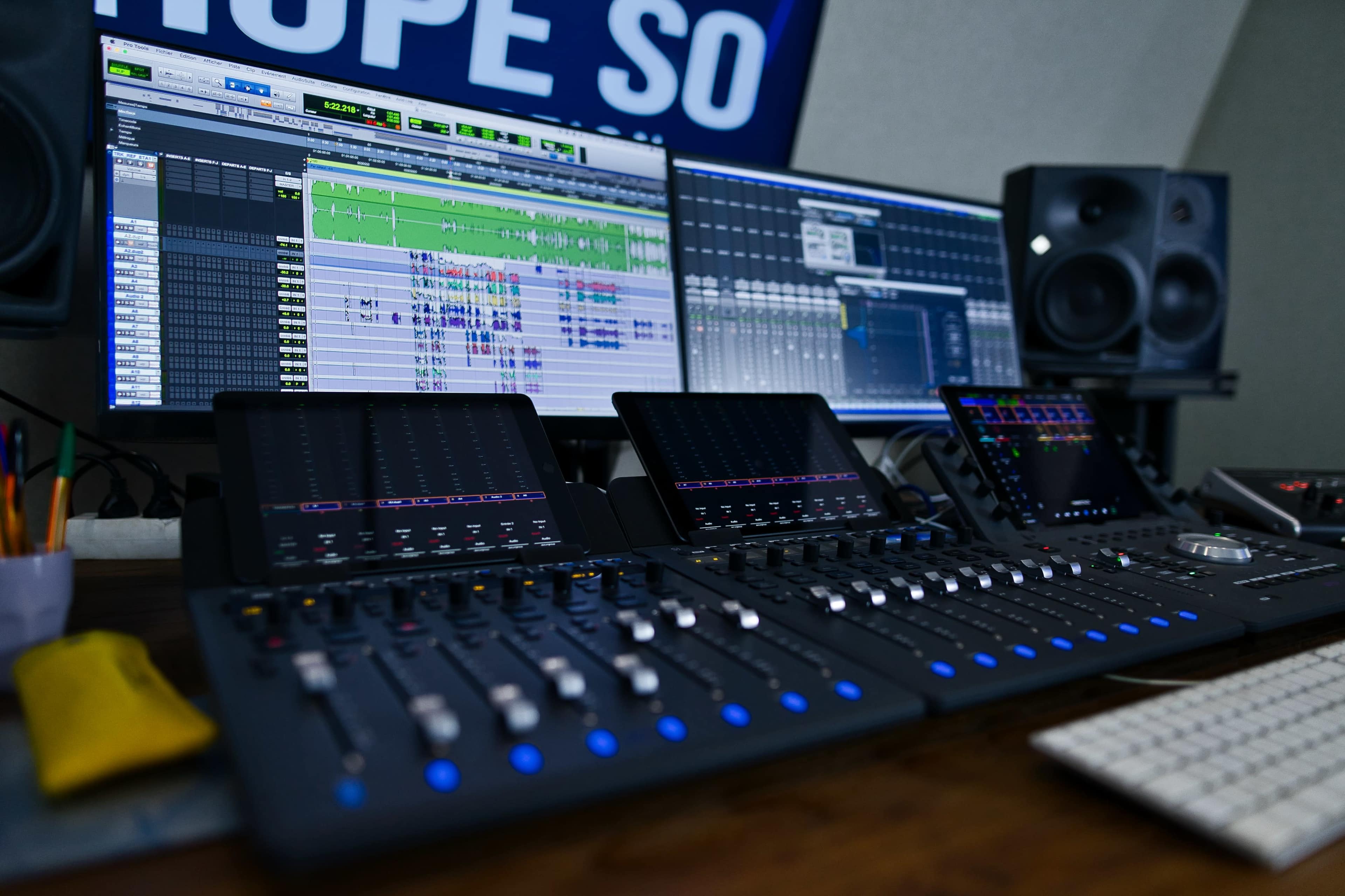 A digital audio workstation with two large monitors displaying audio editing software, a control surface with sliders and buttons, and studio speakers on either side.