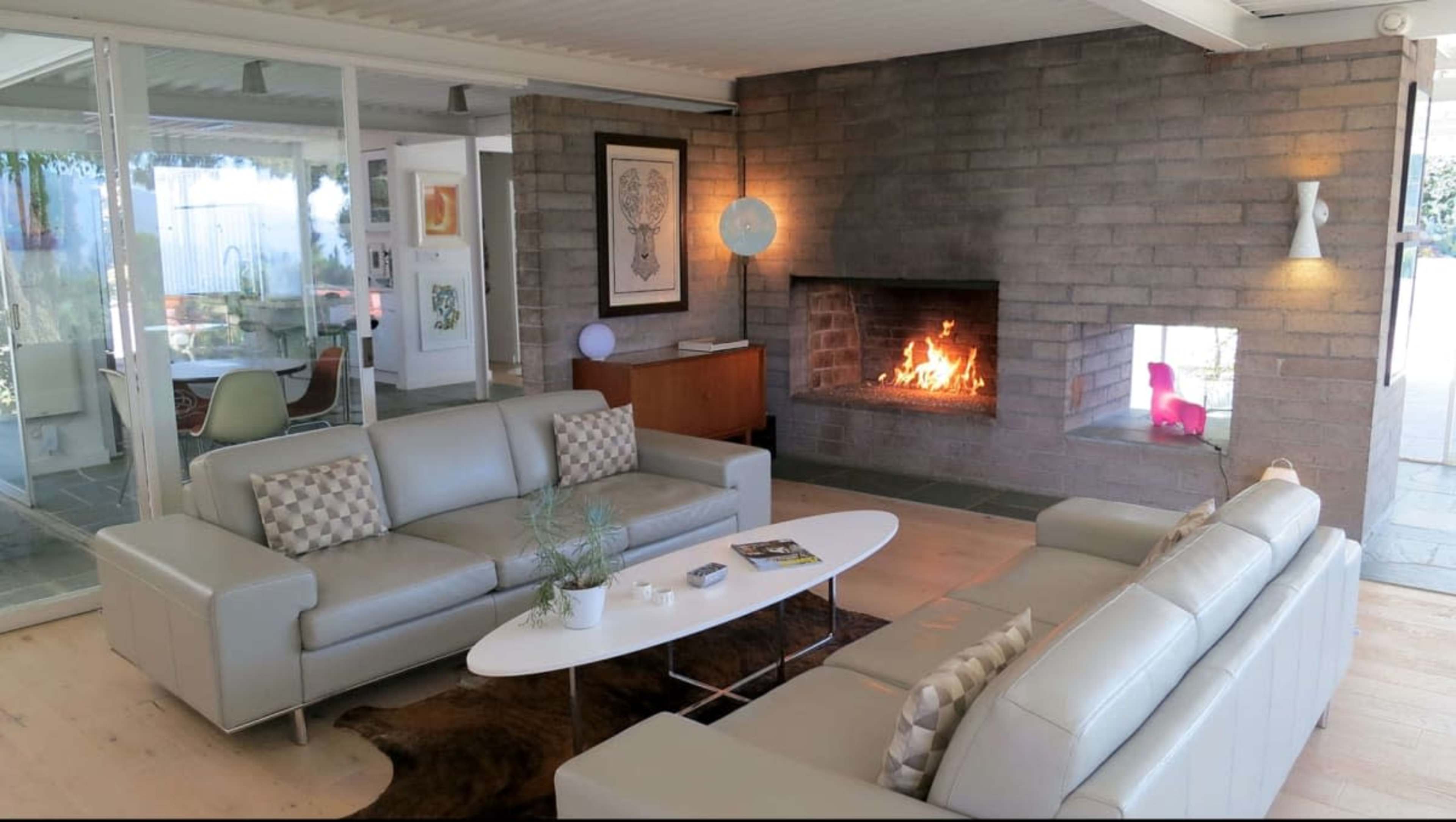 A modern living room features two light-colored sofas facing a central coffee table, with a fireplace and large glass windows showcasing an outdoor view.