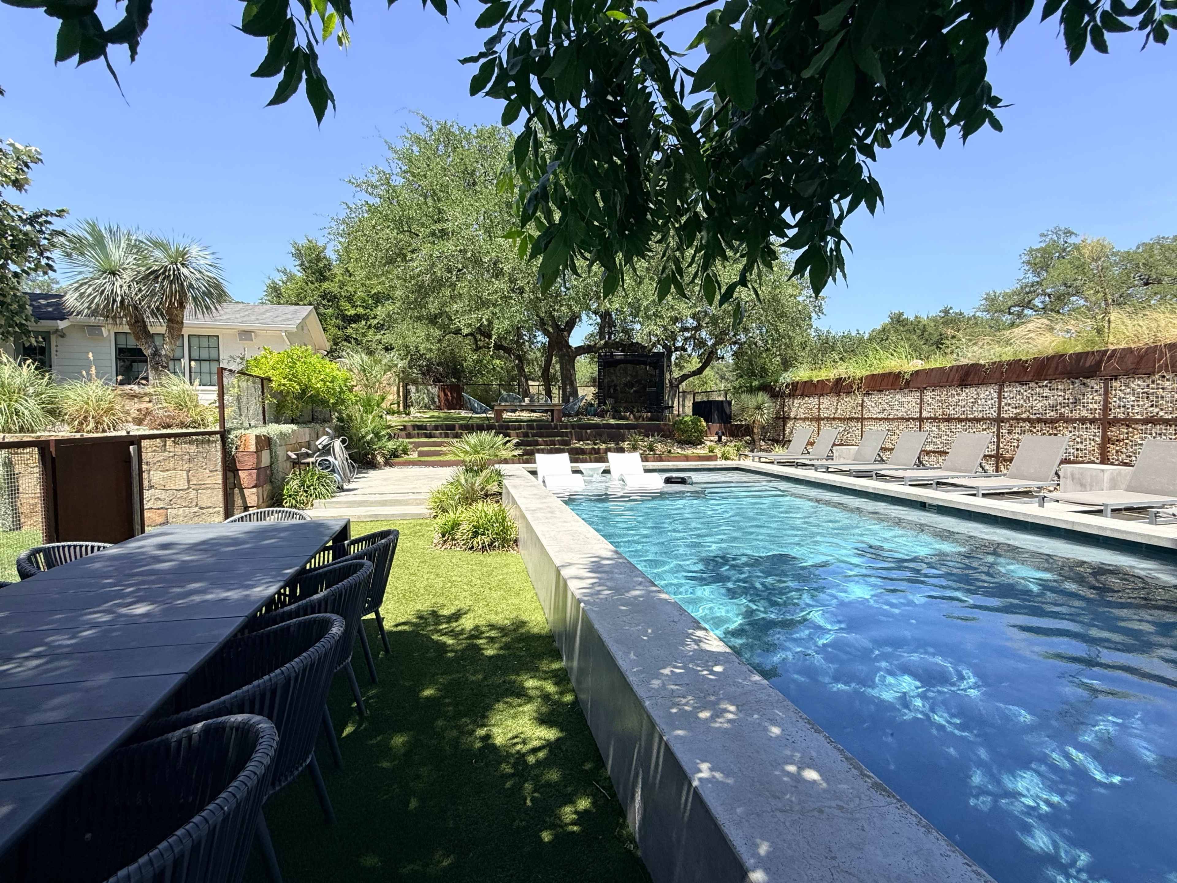 The image shows a modern swimming pool surrounded by lounge chairs and a dining table, with stone pathways and lush greenery in a sunny outdoor setting.