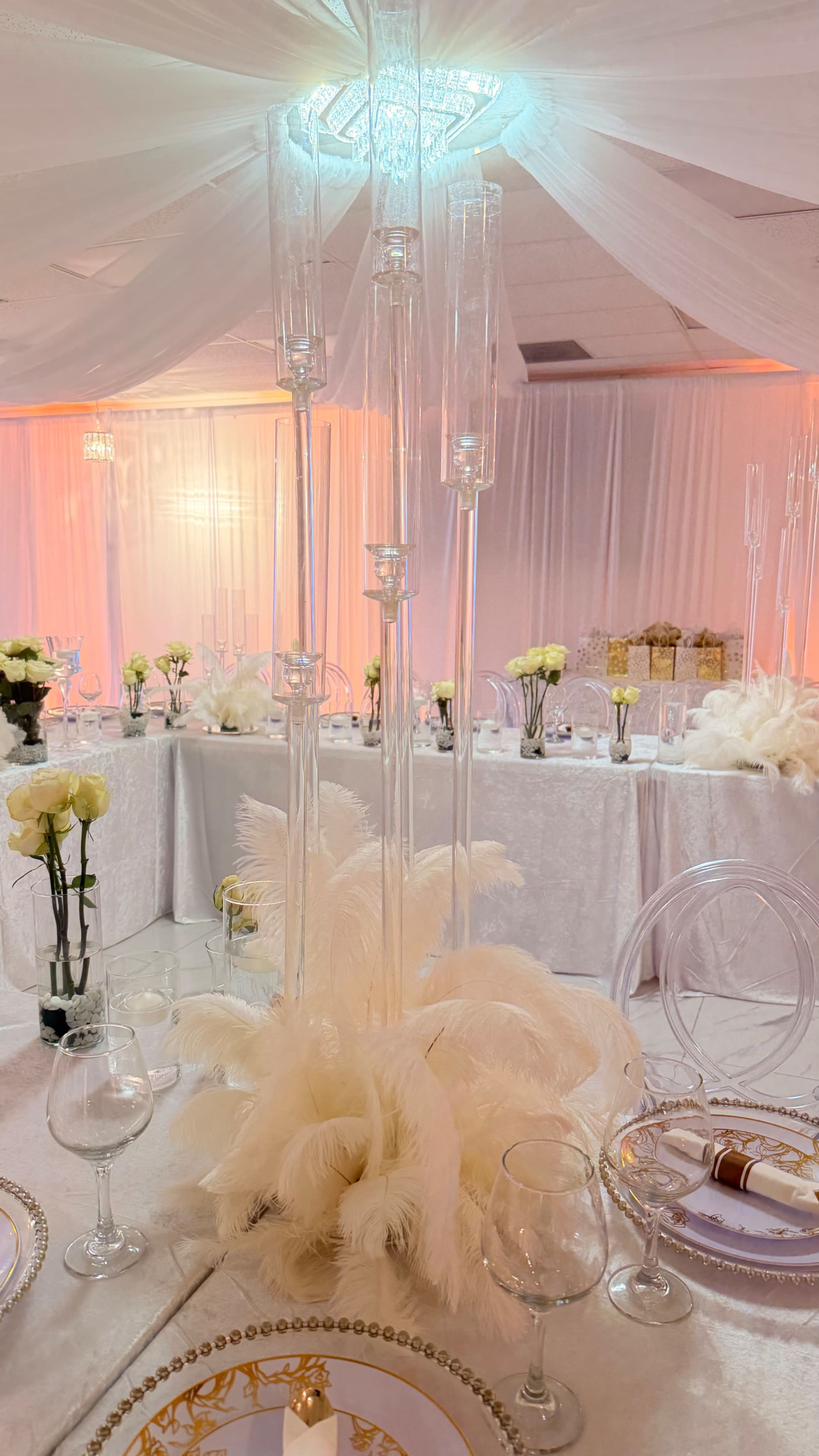 The image shows a elegantly decorated banquet hall with white drapery, glass candle holders, and floral centerpieces on each table.