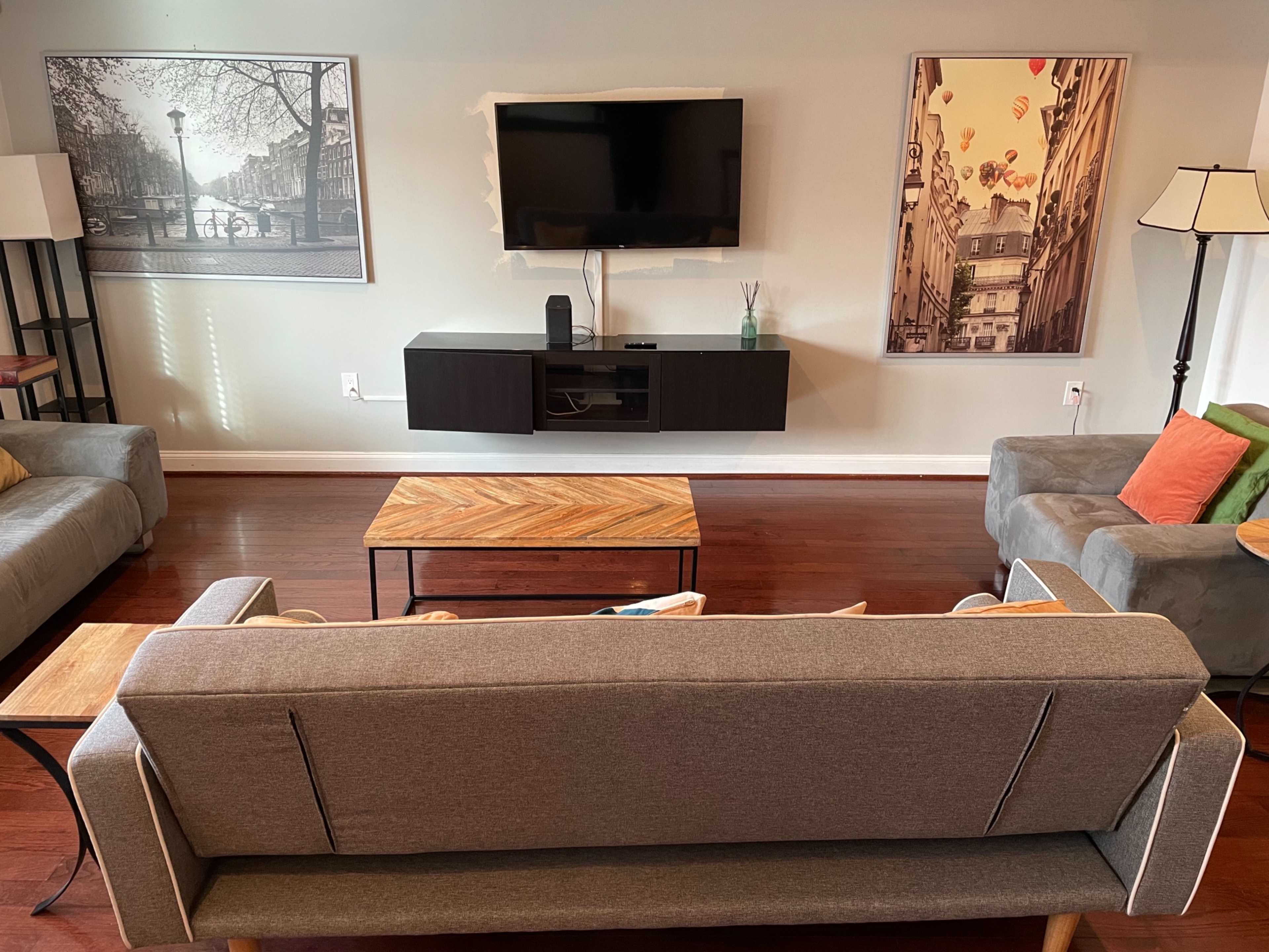 The living room features two gray sofas facing a wooden coffee table, with a television mounted on the wall above a black media console, and artwork displayed on the walls.