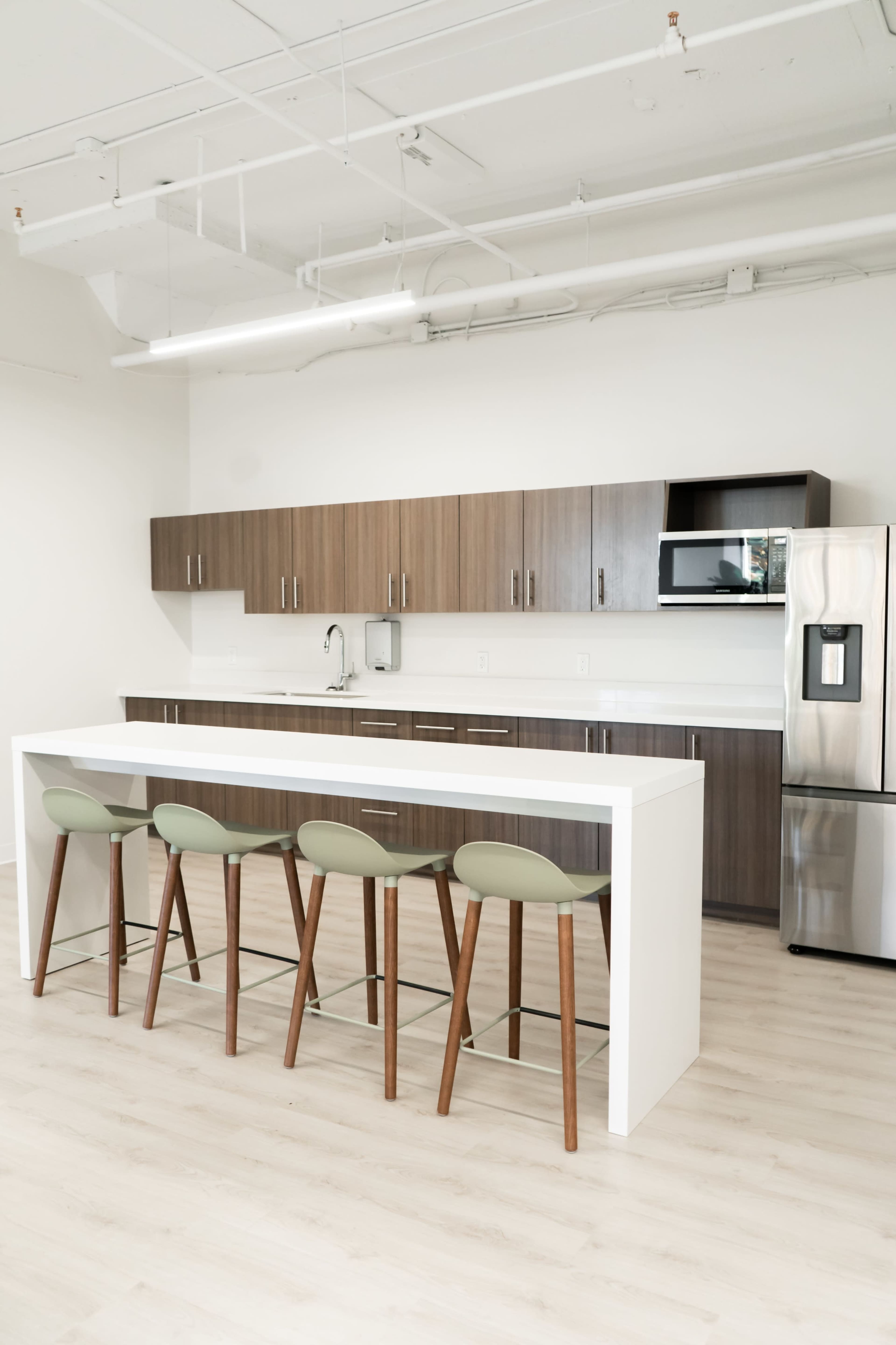 A modern kitchen features a long white countertop with four green stools, dark wood cabinetry, and stainless steel appliances.