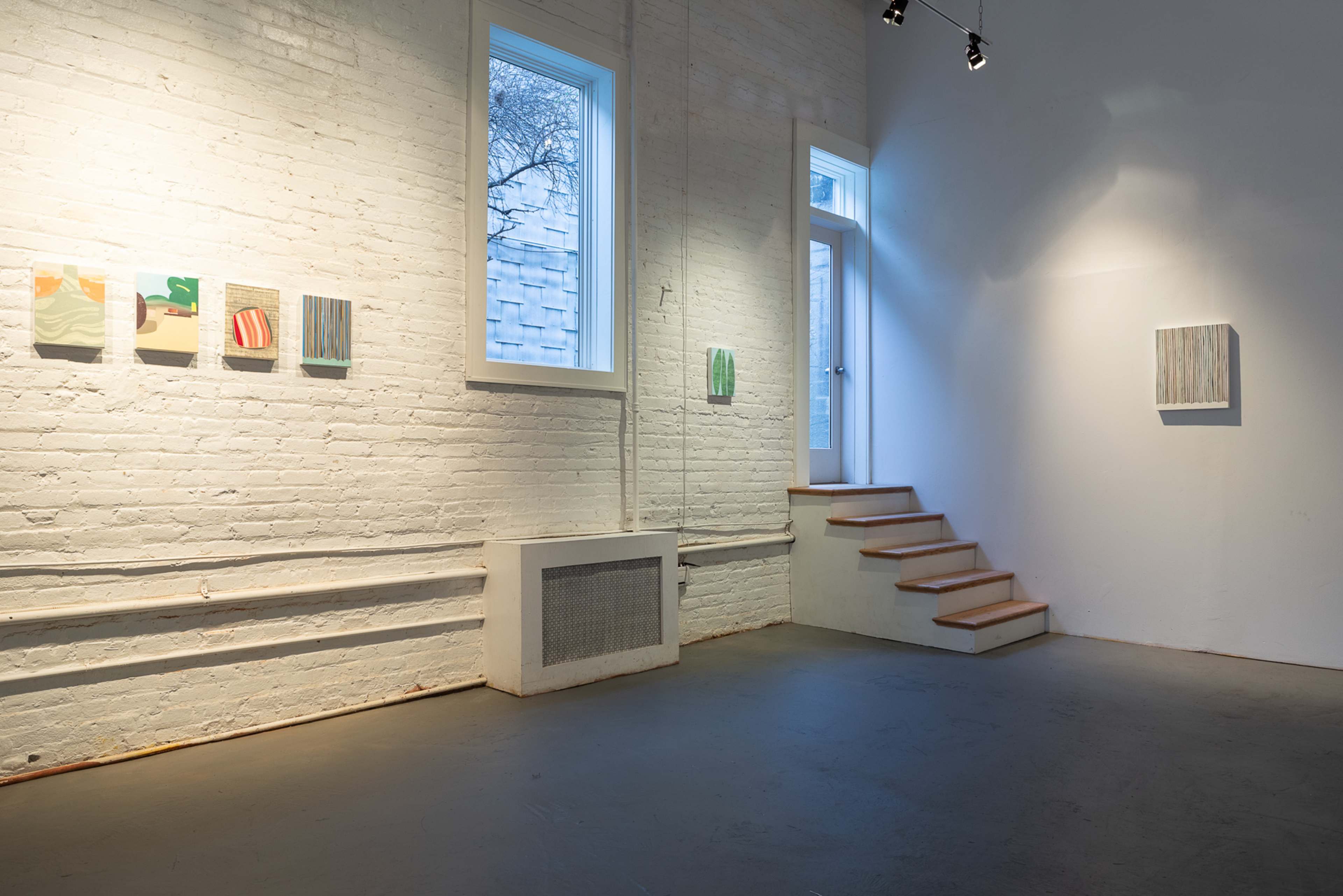 The image shows a minimalist art gallery with whitewashed brick walls, showcasing a selection of paintings on one wall and a staircase leading to another area.