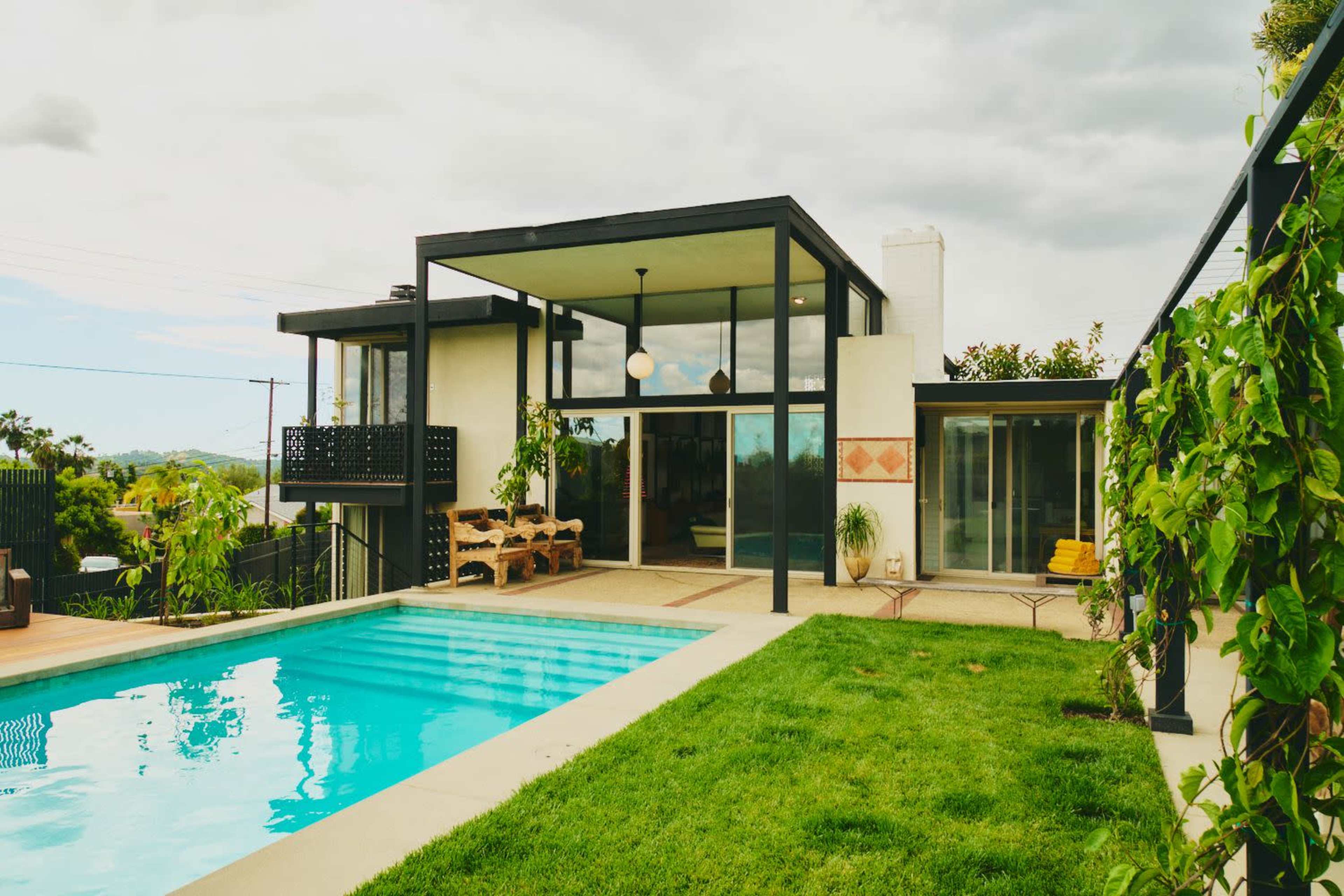 The image shows a modern two-story house with large glass windows, a swimming pool in the foreground, and a well-maintained lawn.