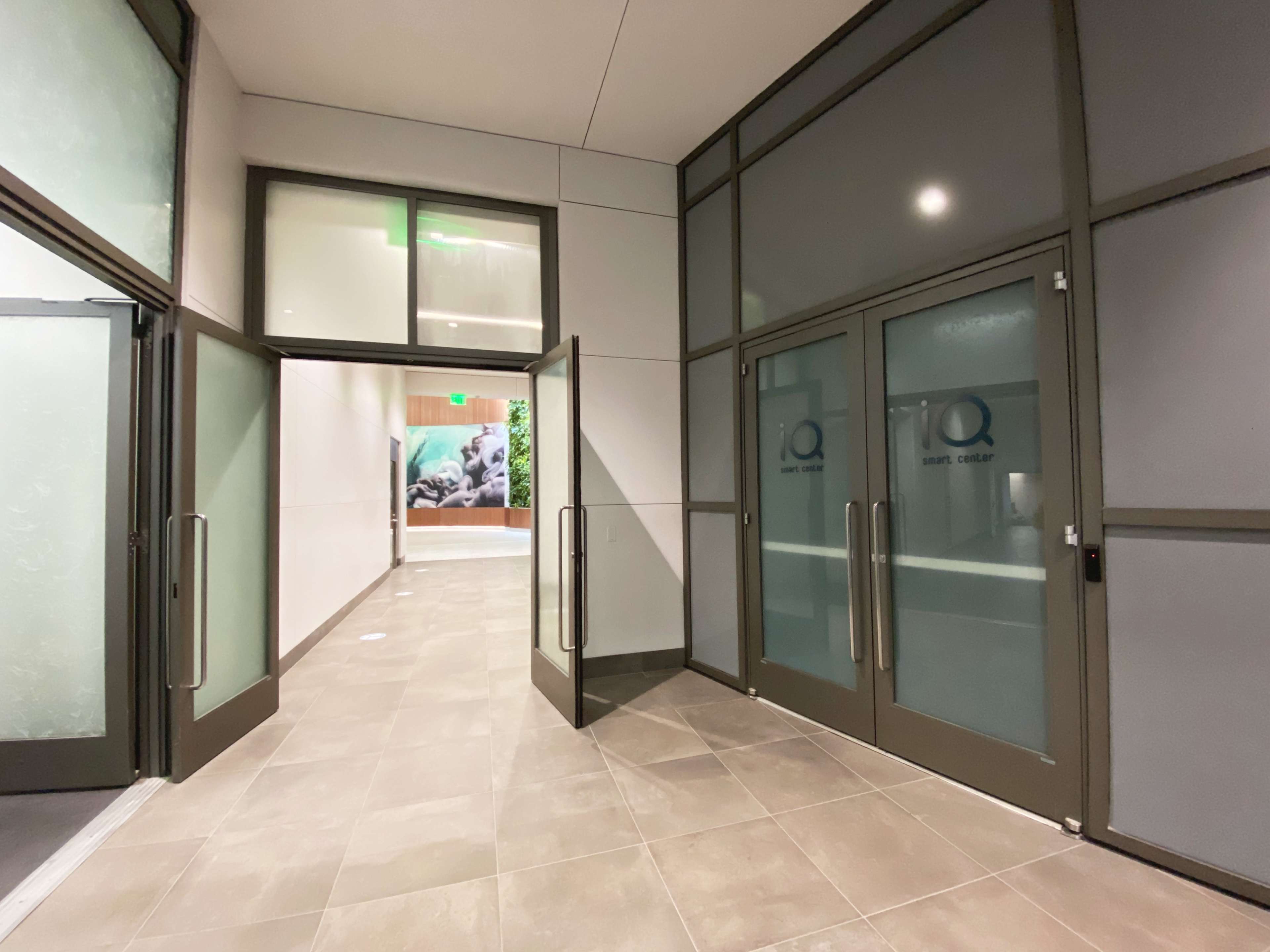 A hallway with glass doors on either side and a tiled floor, leading to another room with greenery visible in the background.