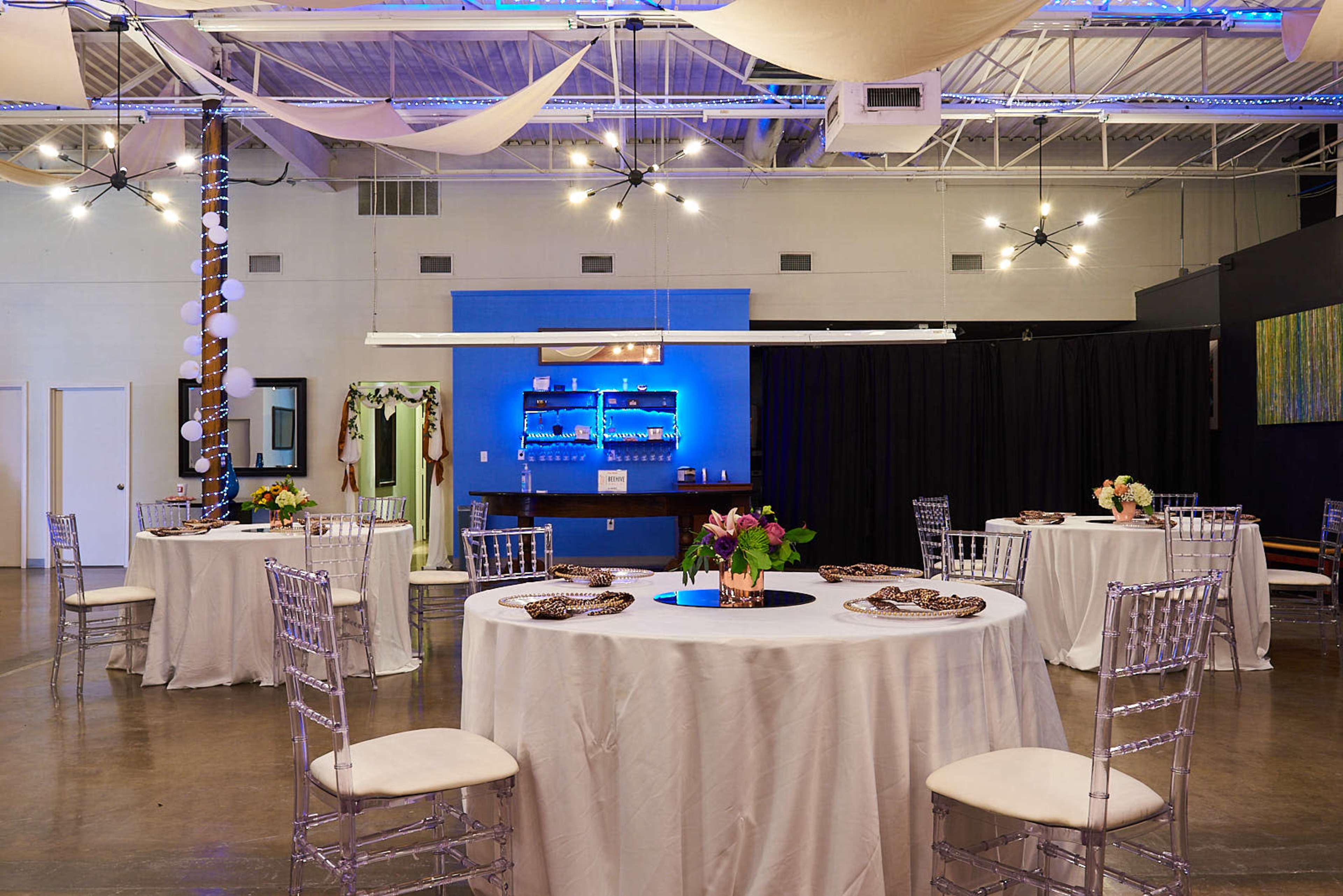 A banquet or event space with round tables set with tablecloths, centered plates, and floral arrangements, alongside a bar area illuminated with blue lighting.