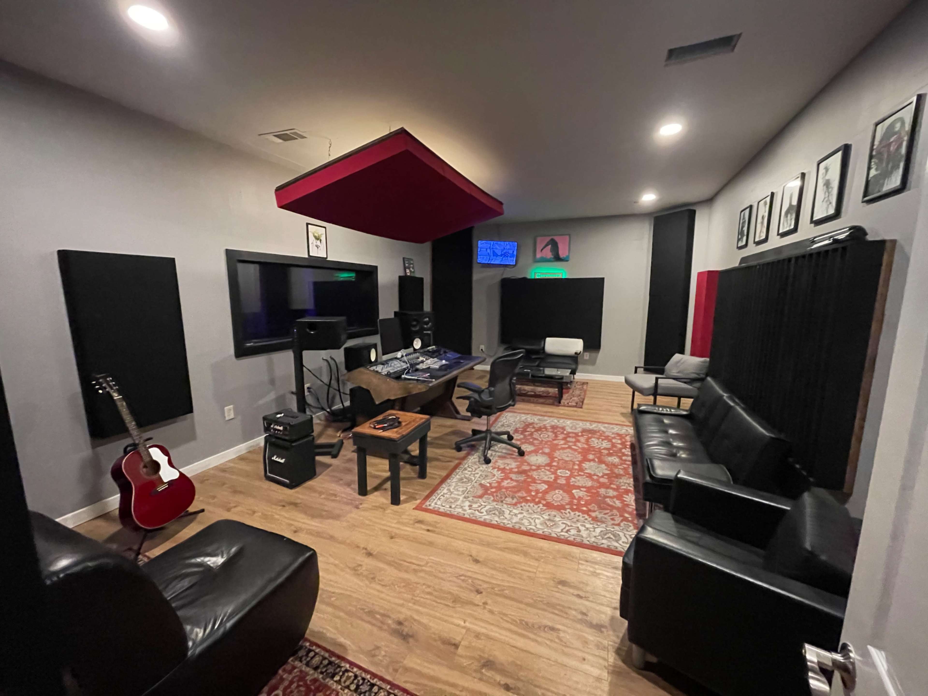 The image shows a modern music recording studio with guitars, soundproofing panels, and a control desk surrounded by seating.
