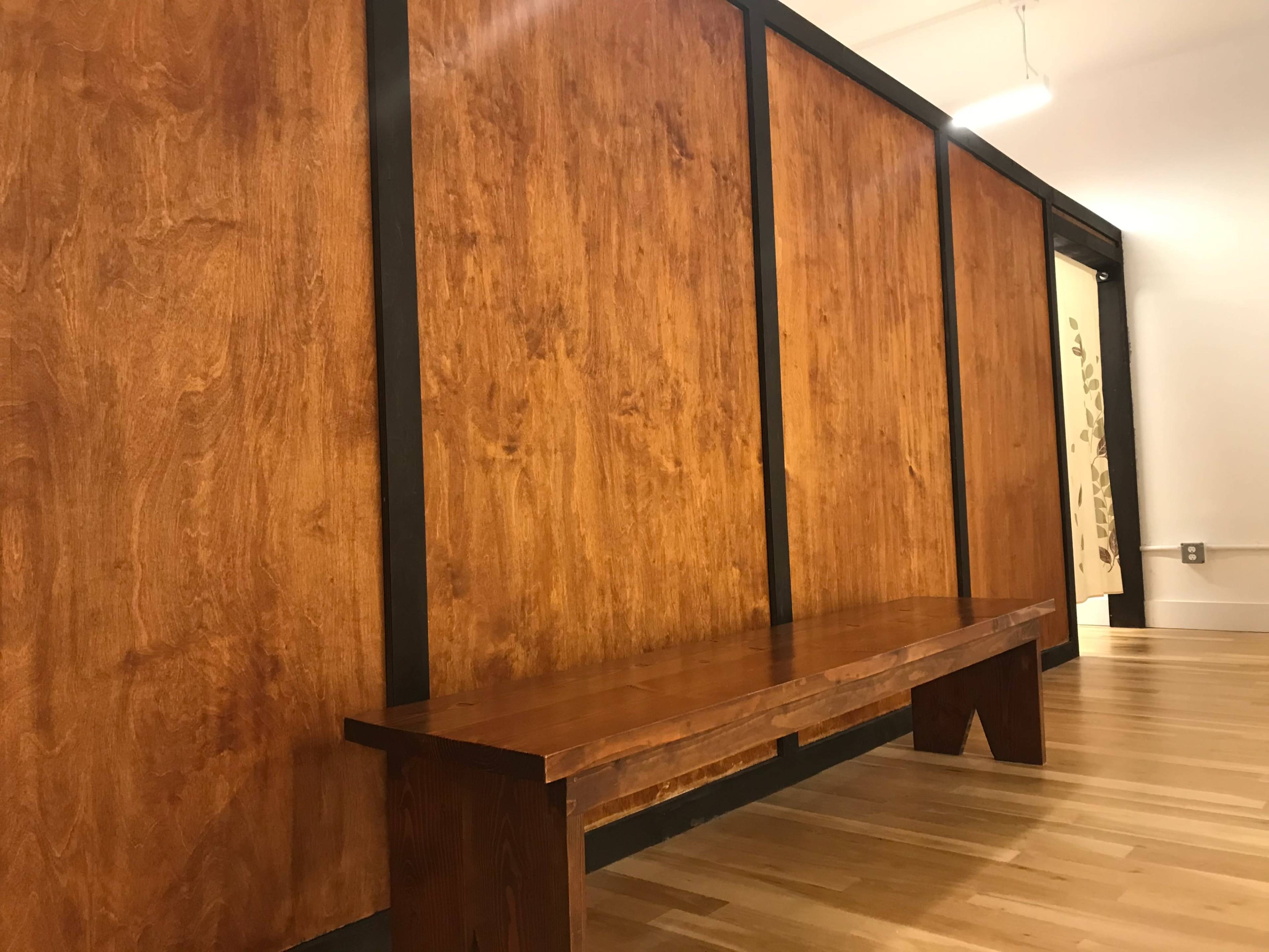 The image shows a wooden bench against a backdrop of large, smooth wooden panels in a well-lit interior space.