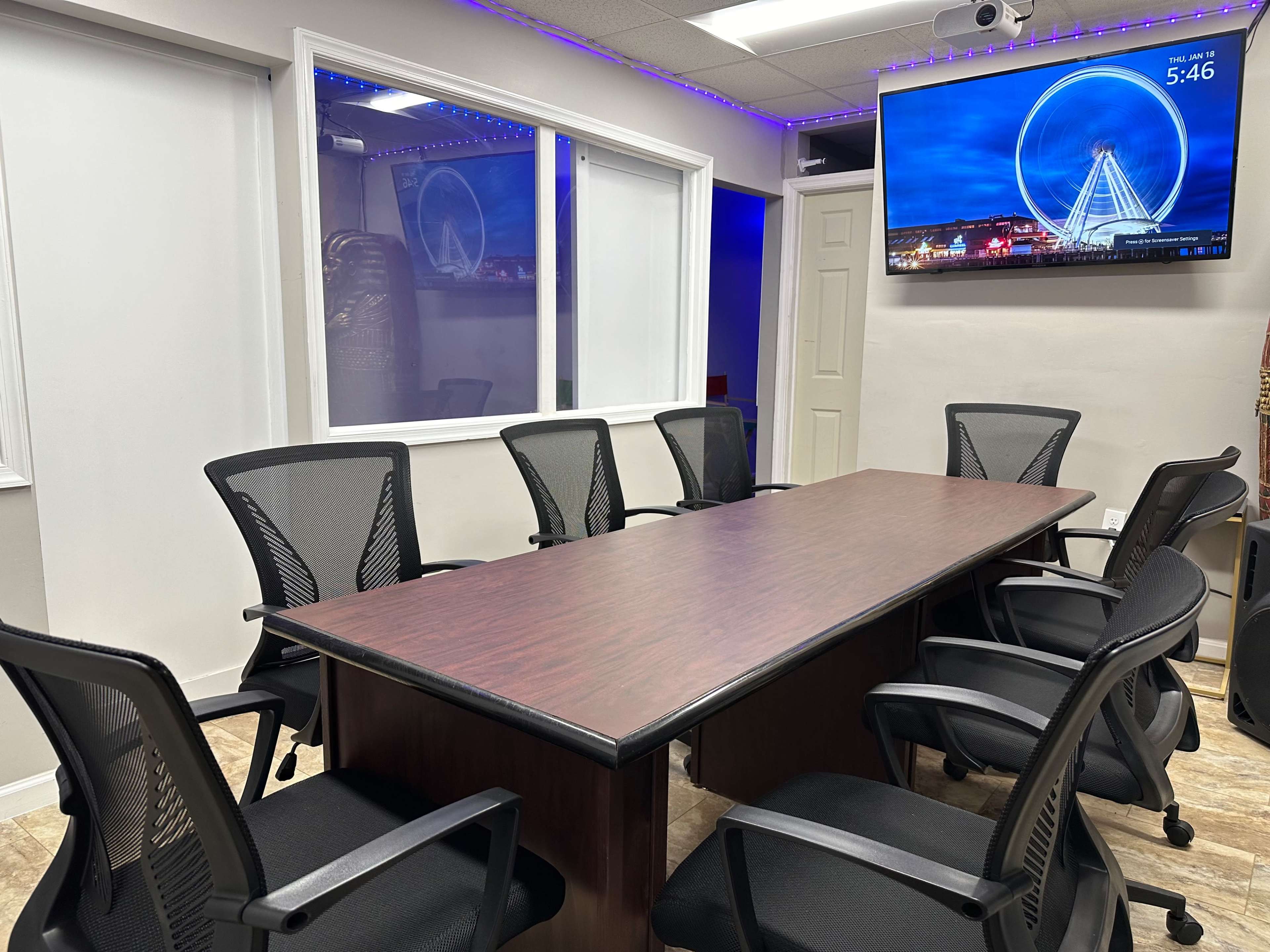 A conference room features a large wooden table surrounded by six black mesh chairs, with a television displaying a Ferris wheel image on the wall.