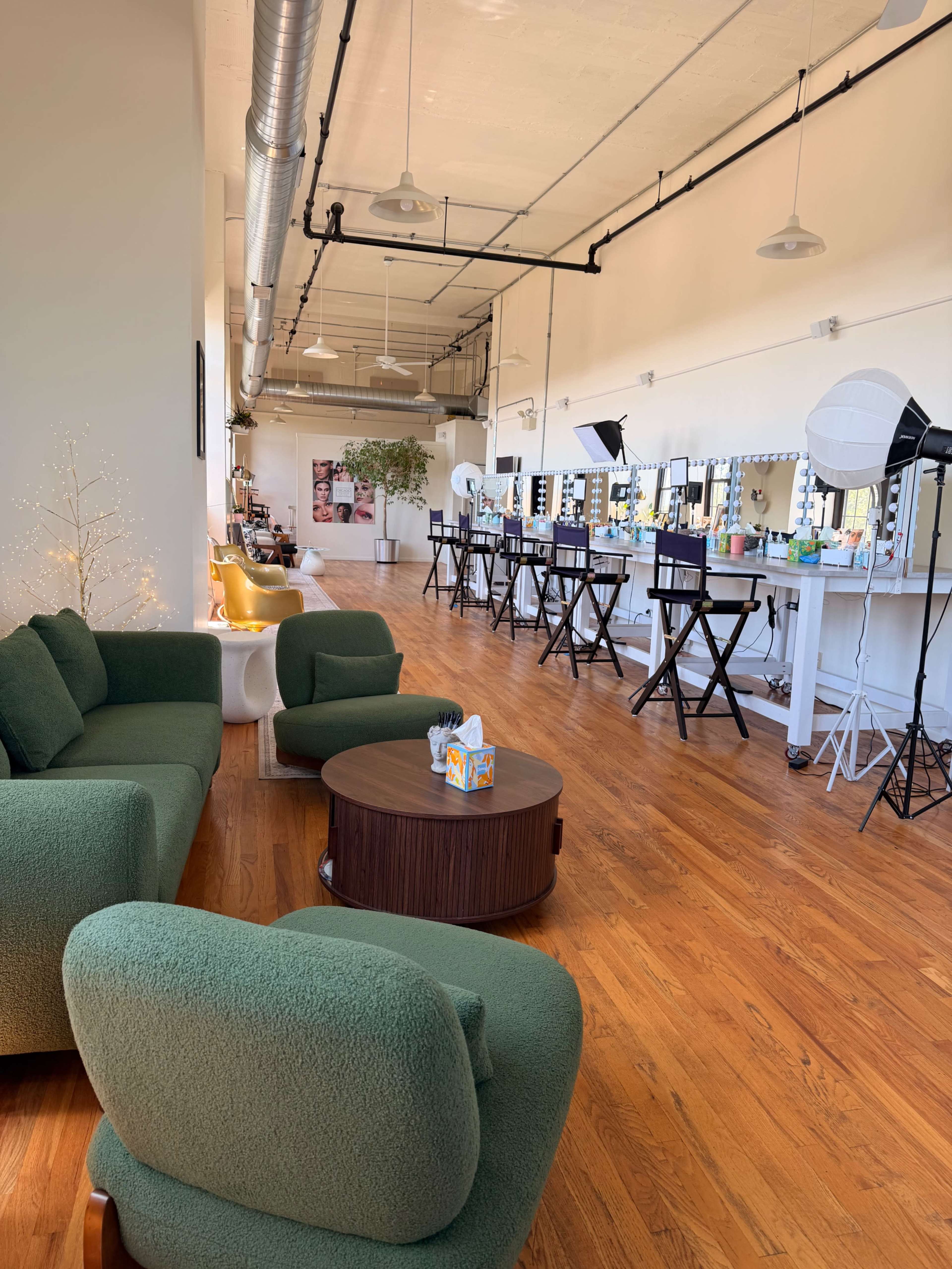 The image shows a spacious beauty studio with green seating, wooden flooring, and several makeup stations equipped with large mirrors and lighting.