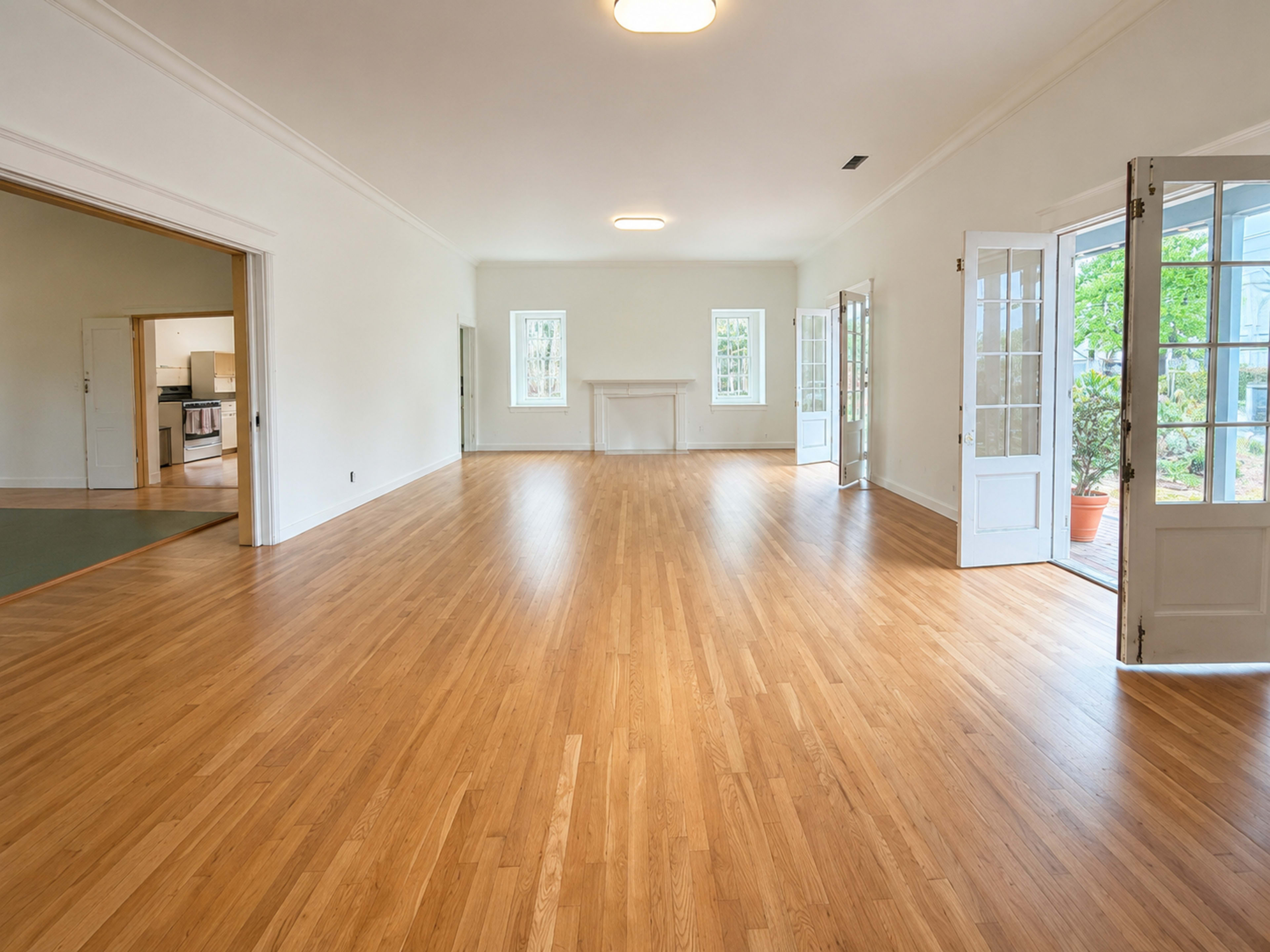 The image shows a spacious, empty room with hardwood floors and large windows, featuring double doors that open to an outdoor area.