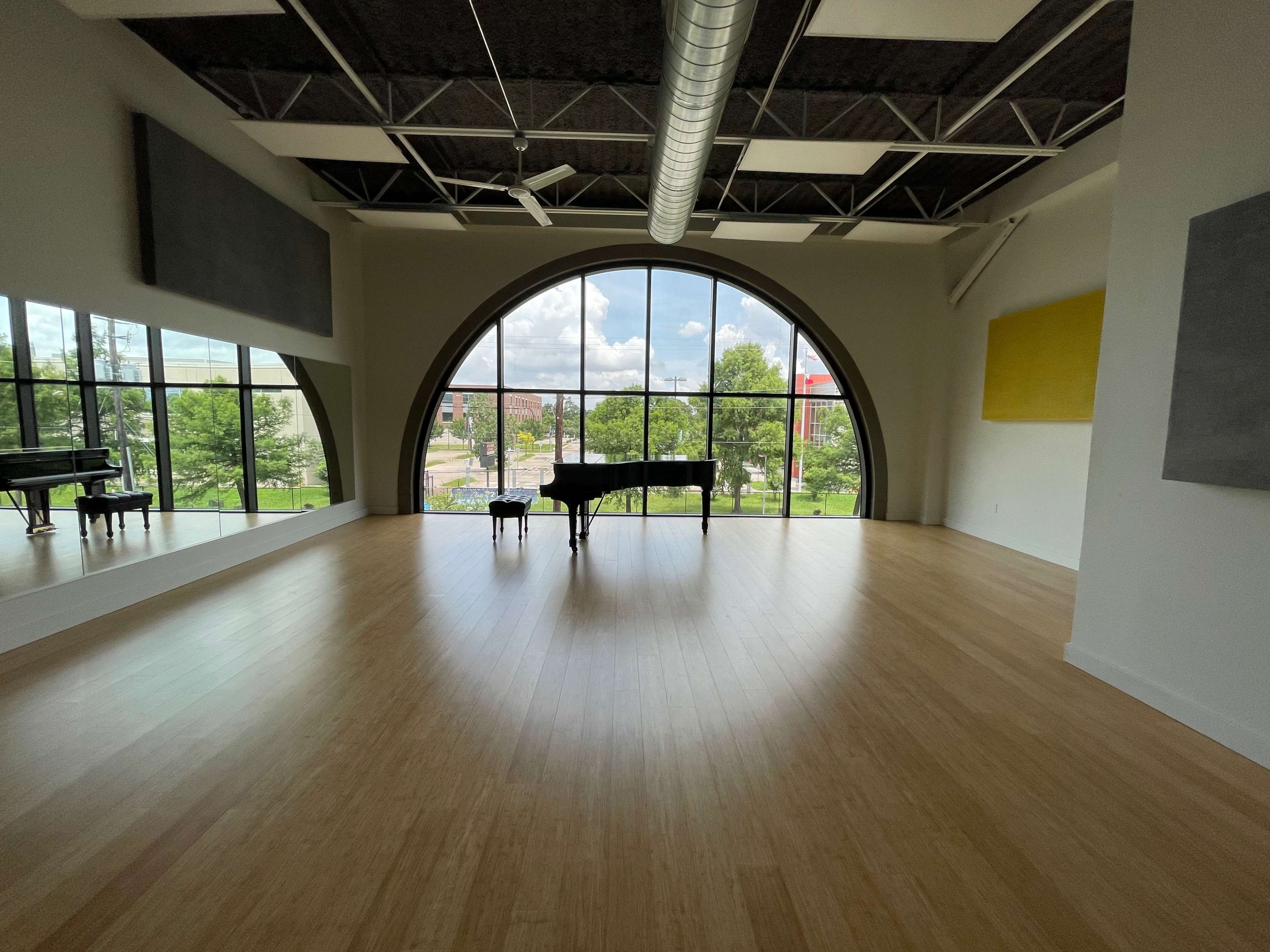 A spacious music studio features a grand piano positioned in front of a large arched window, overlooking a tree-lined street.