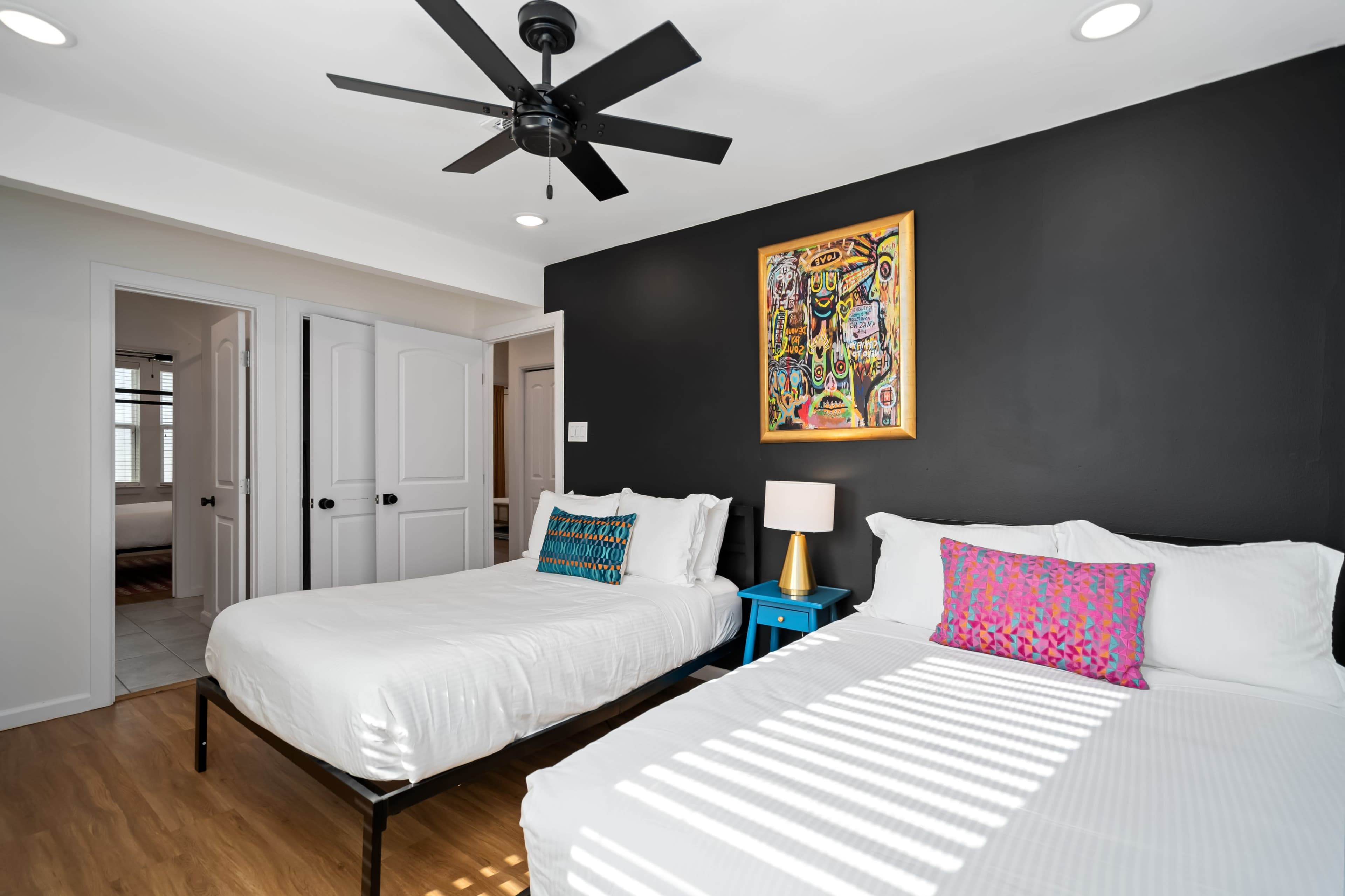 A modern bedroom features two beds with white linens, a colorful decorative pillow on one, a nightstand with a lamp, and a piece of wall art above one bed, against a dark accent wall.