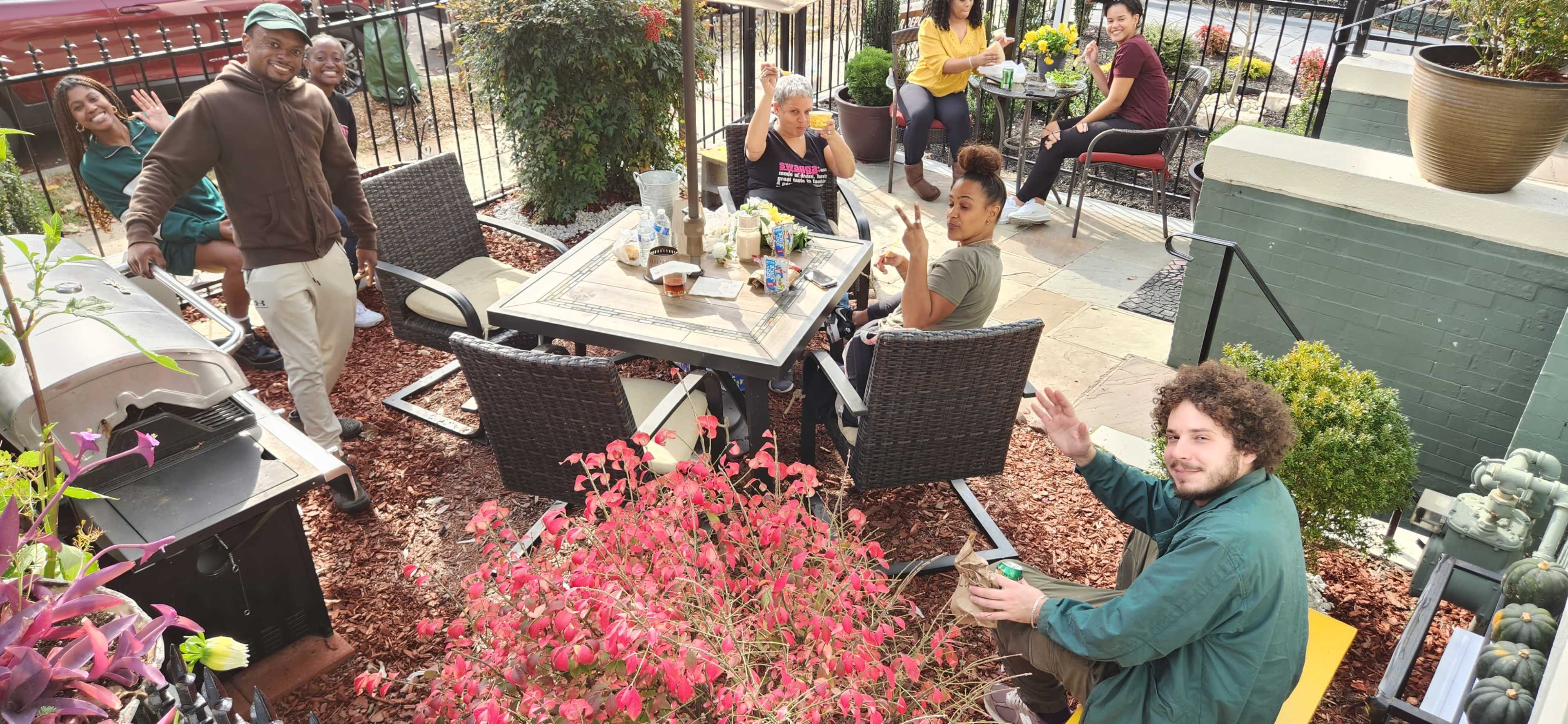 A group of people are gathered around a table on an outdoor patio, enjoying food and drinks while smiling and waving at the camera.