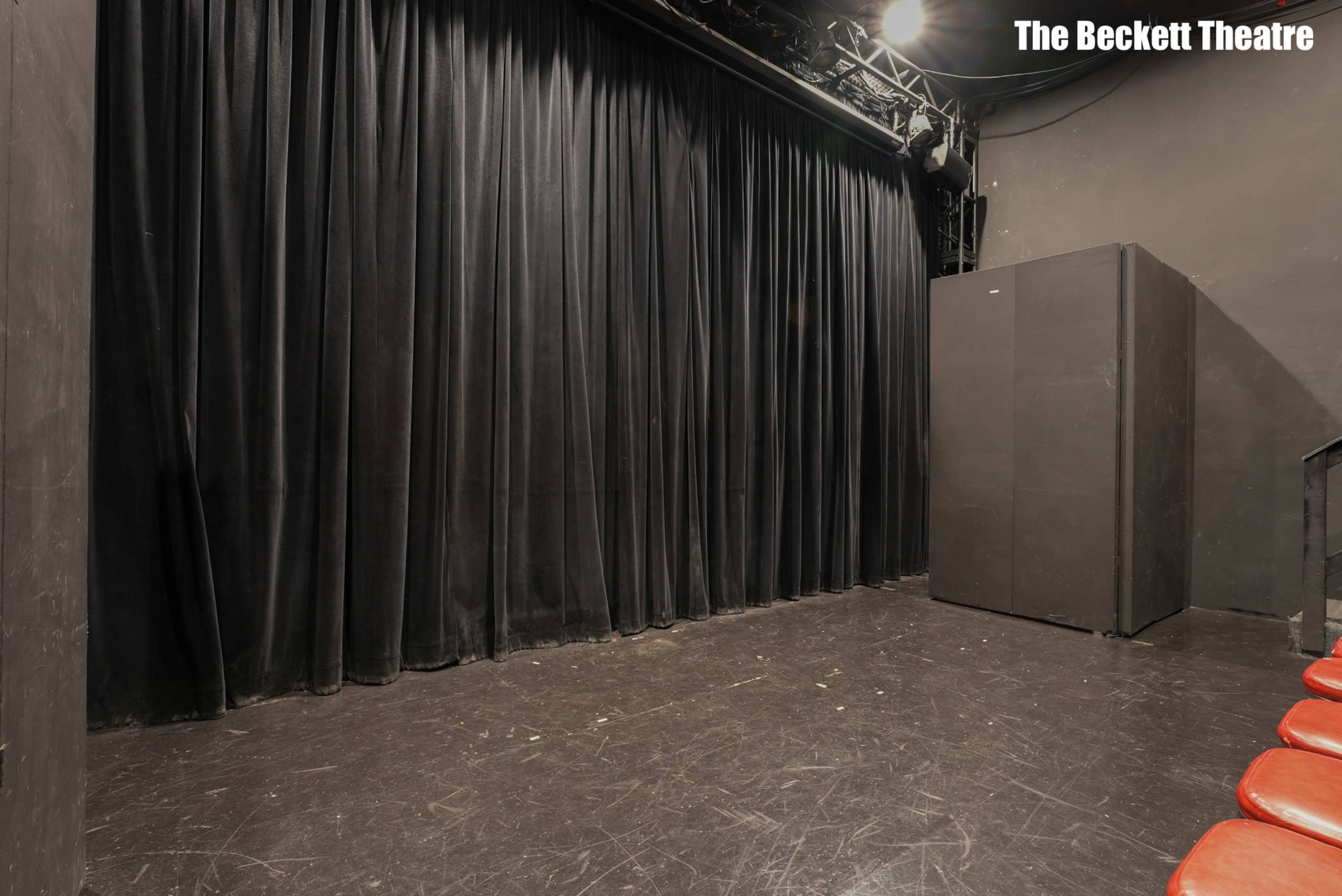The image shows a dimly lit stage with black curtains and a large dark storage unit in the corner.