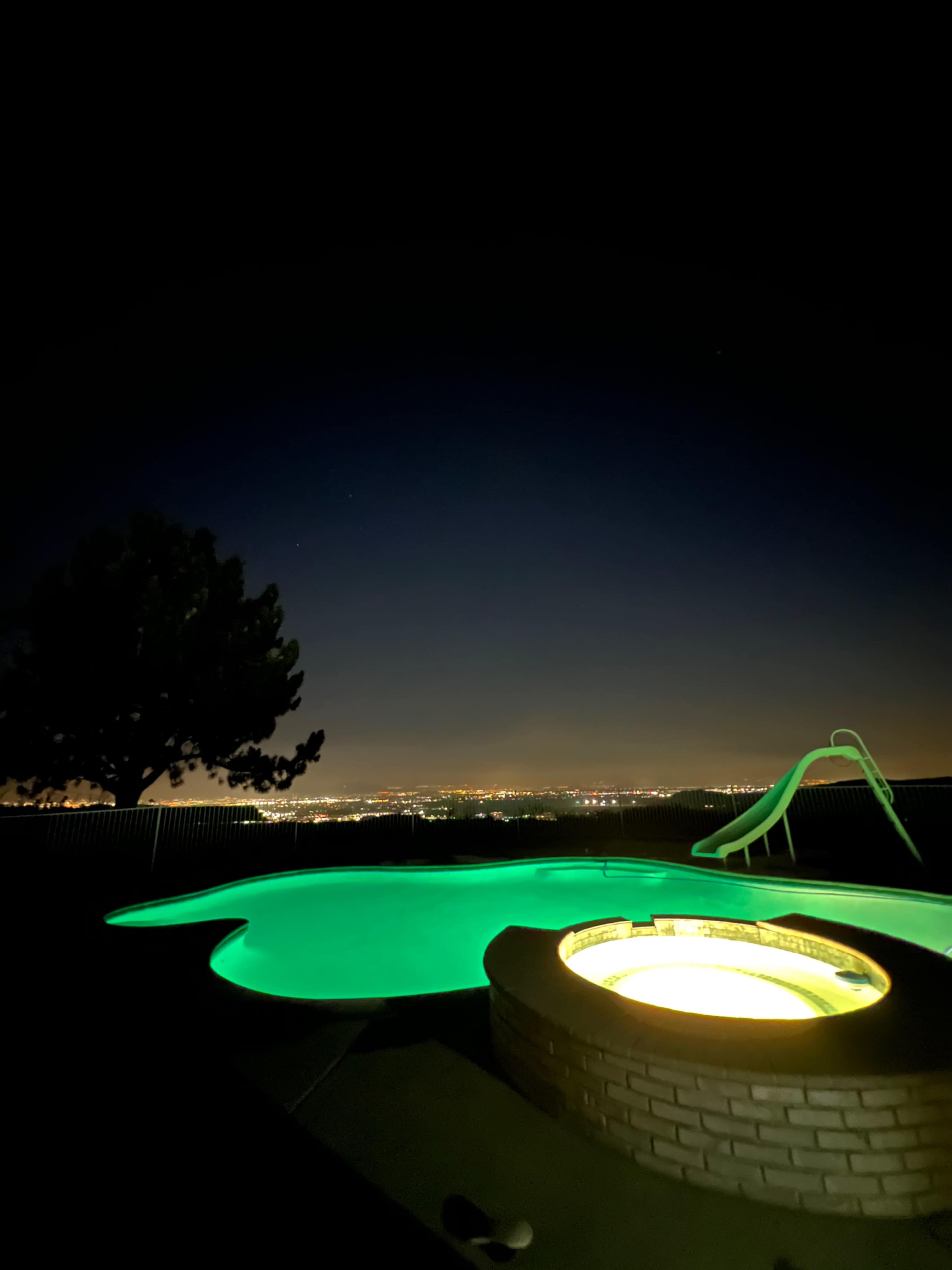 A brightly lit hot tub beside a dark swimming pool, set against a city skyline at night.
