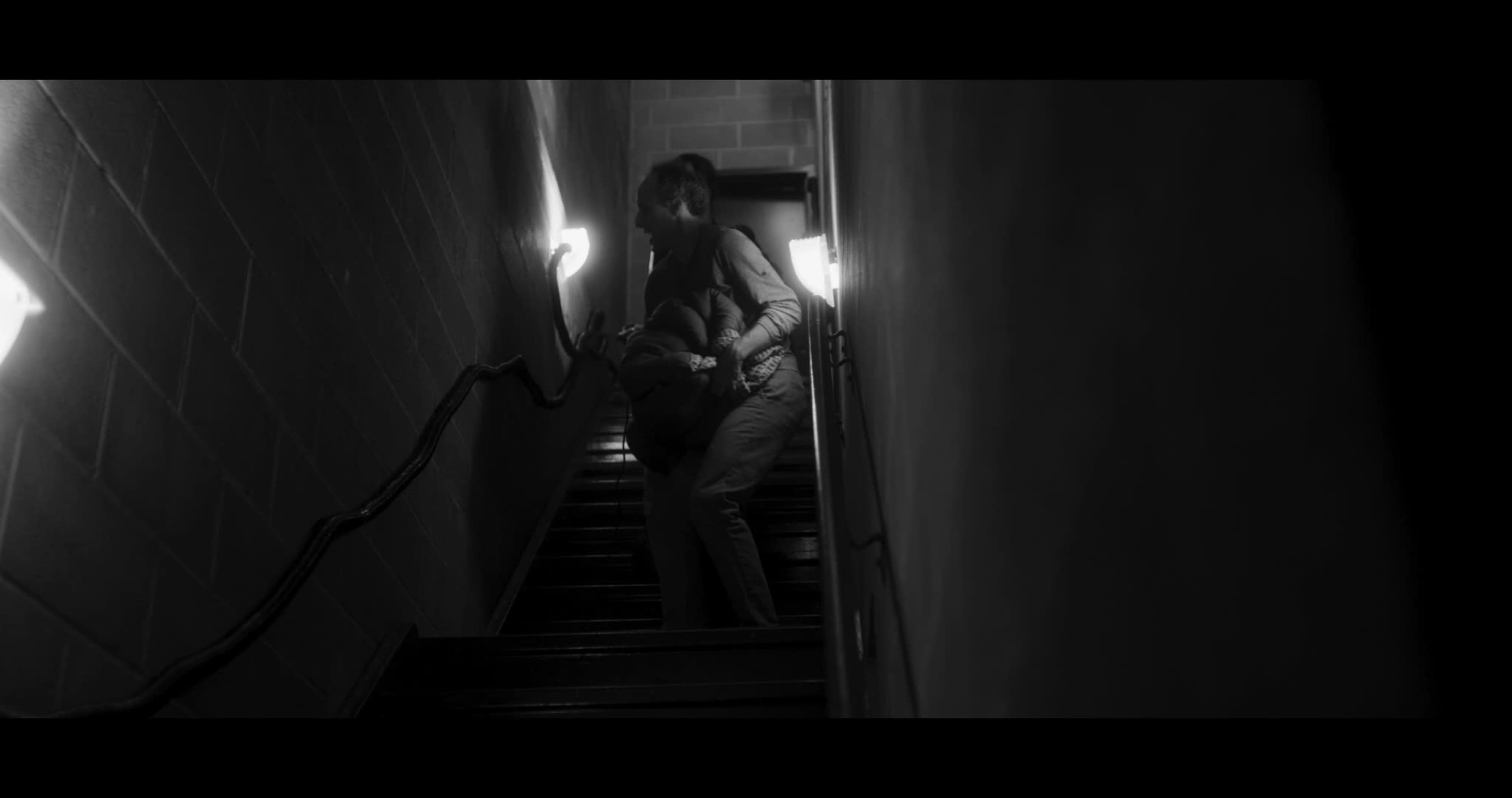 A person lifts another person while climbing a narrow staircase in a dimly lit corridor.