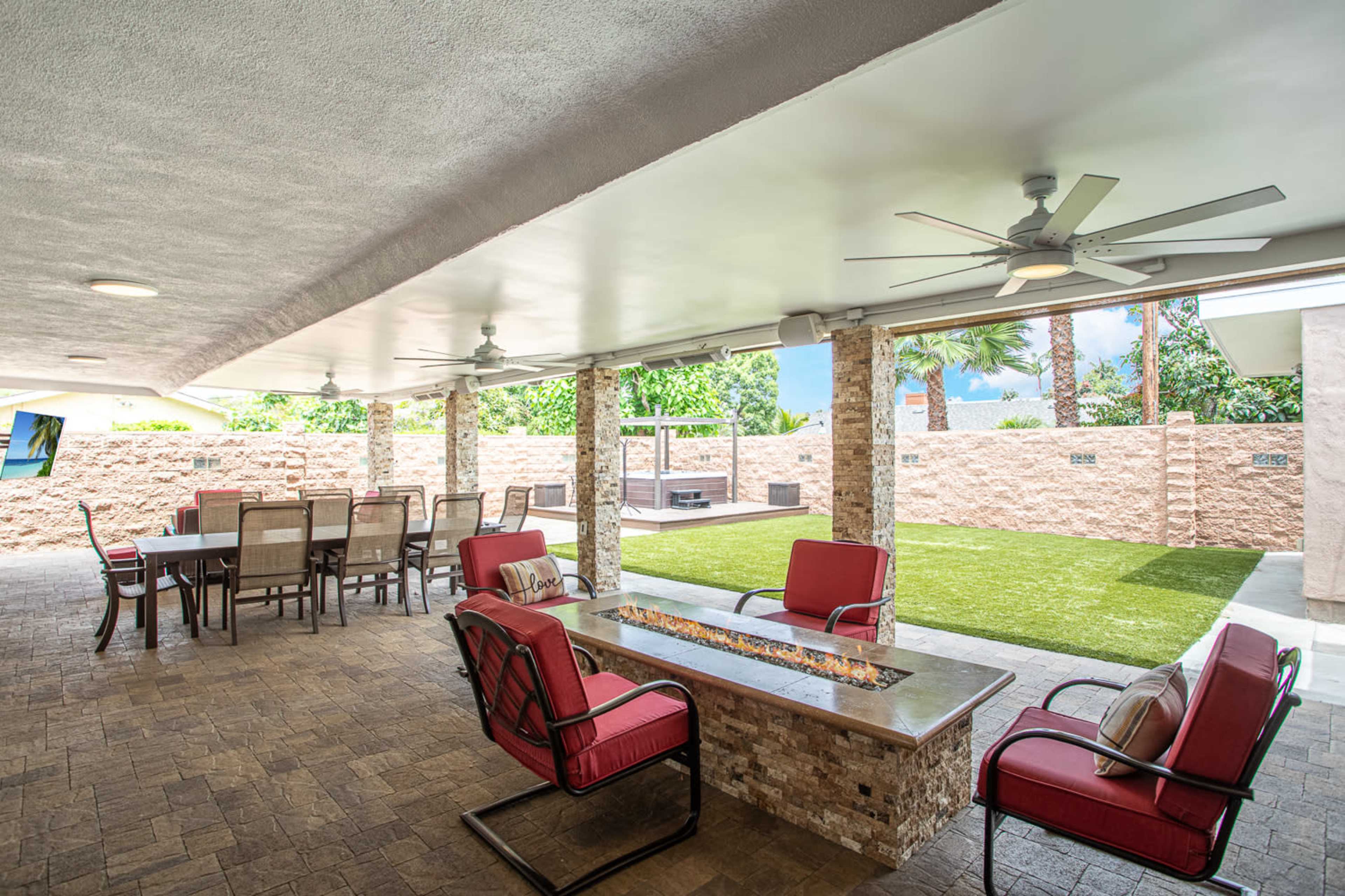 A covered outdoor patio area featuring seating with red cushions, a fire table, a dining table, and artificial grass.