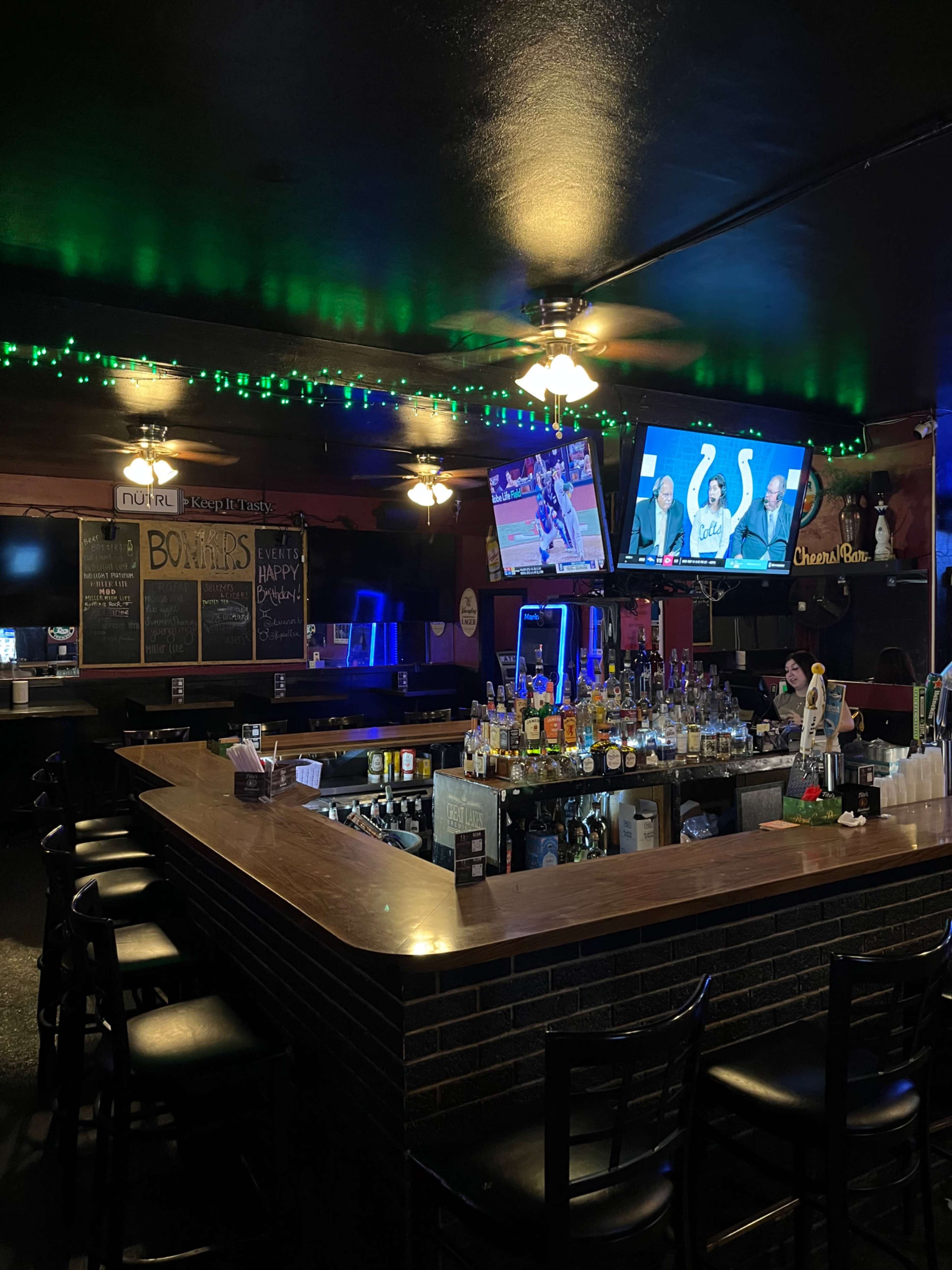 The image shows a dimly lit bar with a wooden bar top, multiple TV screens displaying sports, and a variety of bottles lined up behind the counter.