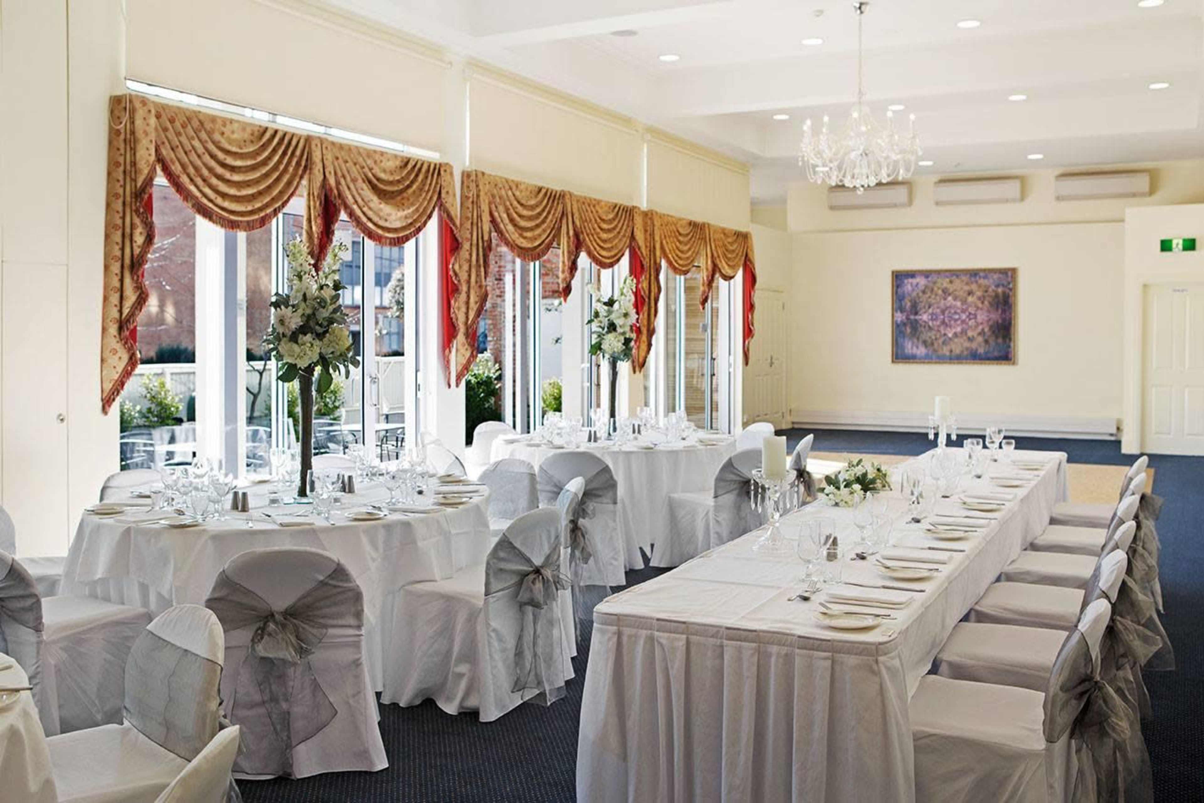 A banquet room is set up for an event with neatly arranged tables covered in white linens and decorated with floral centerpieces near large windows.